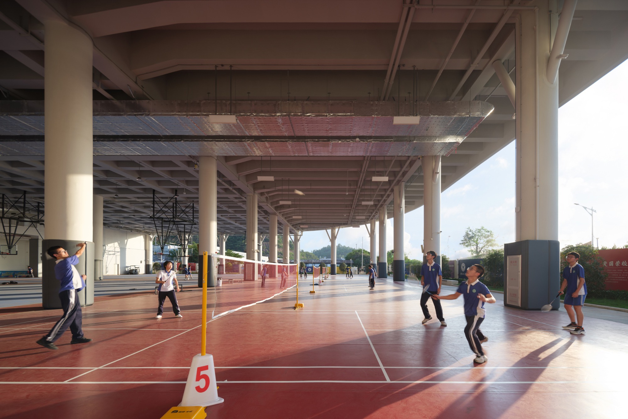 Covered red-floored badminton court beneath the elevated structure, with players, a net on yellow posts and low sun from the right