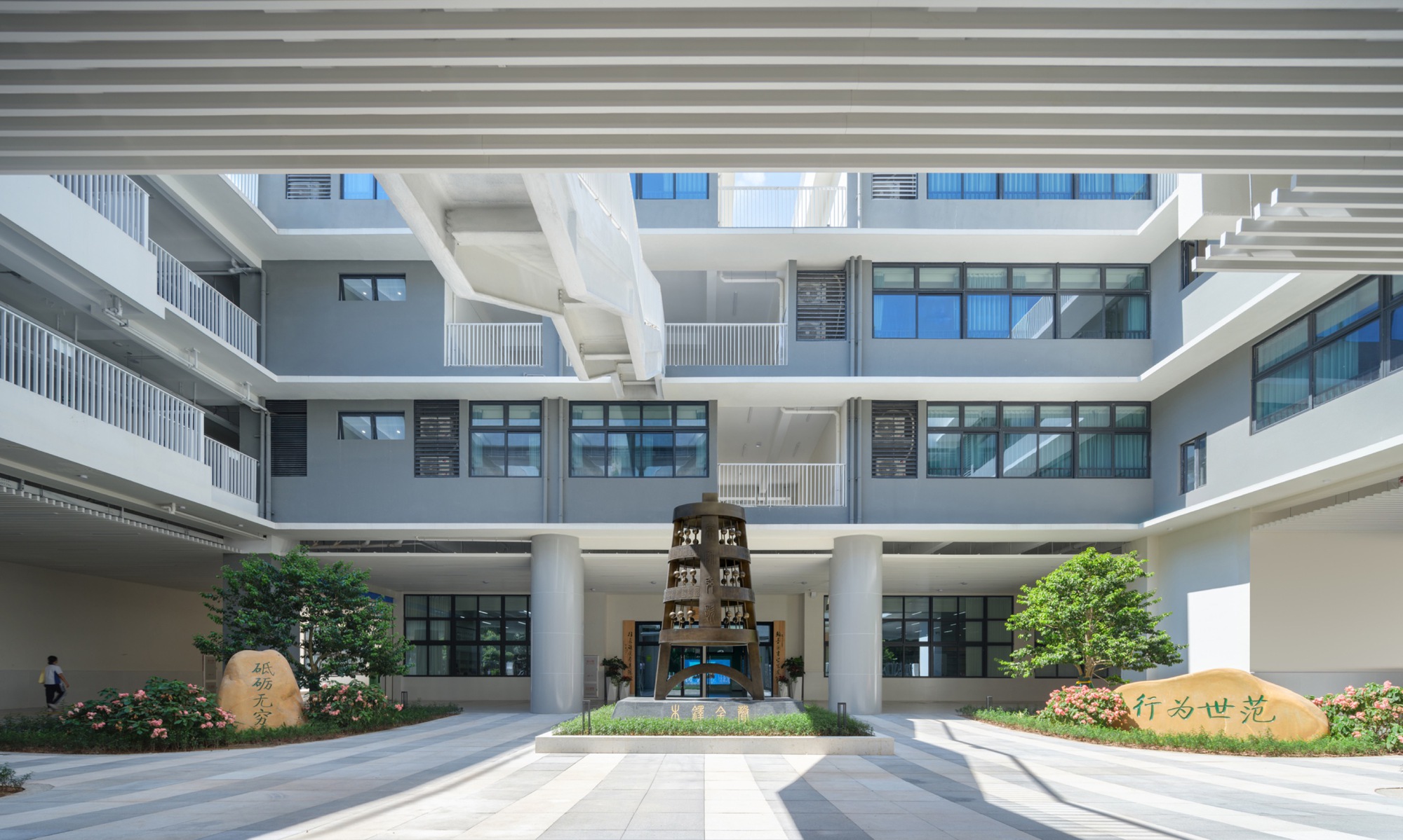 Courtyard with a bronze sculpture between two round columns, inscribed boulders and planted trees below stacked grey balconies in daylight