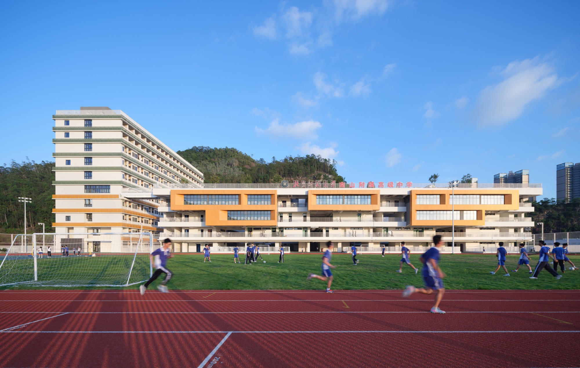 Students running across the red track and grass pitch in front of the white school block with orange bays