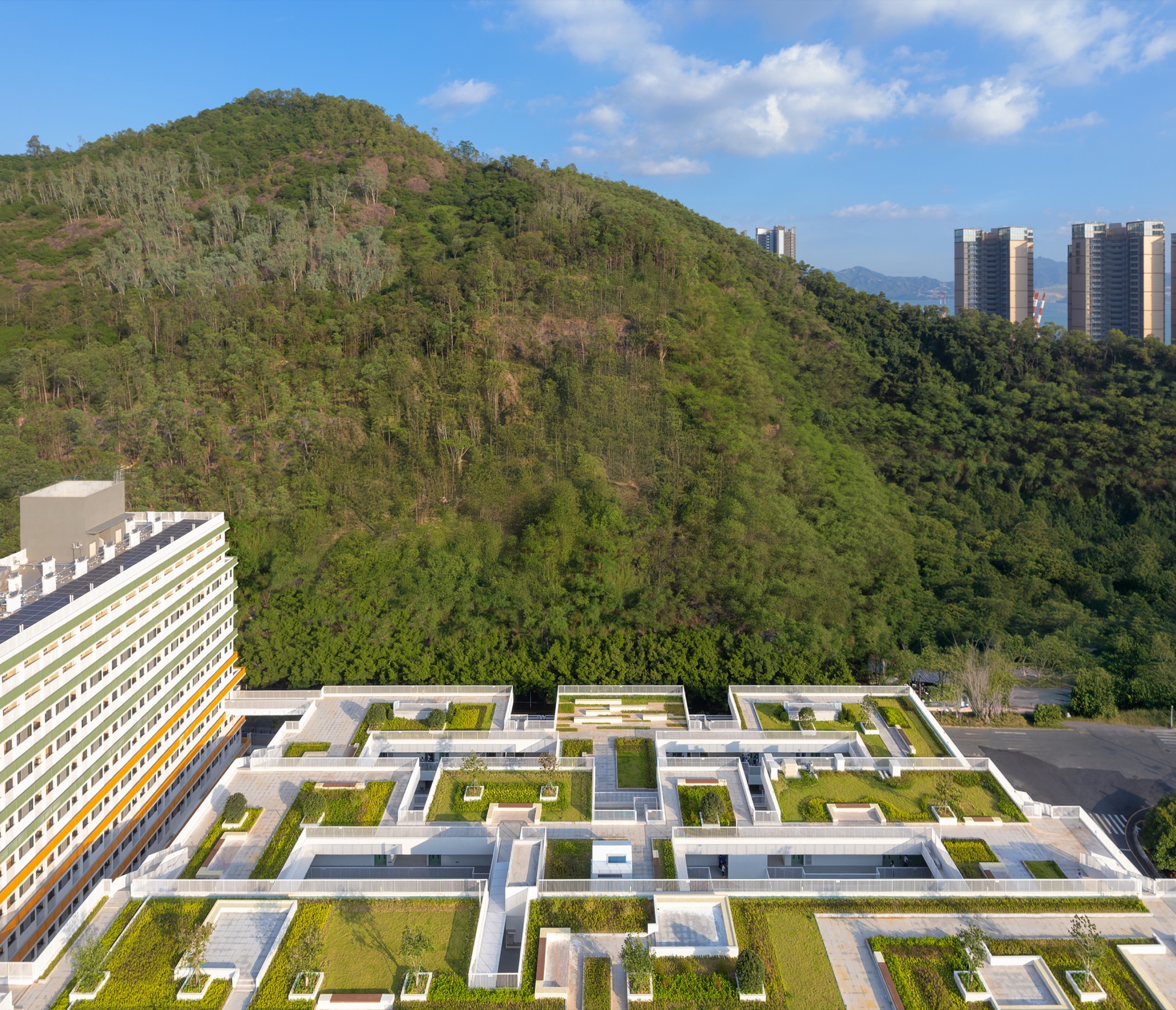 Aerial view of terraced green roof courtyards with white parapets below a forested hillside and distant towers