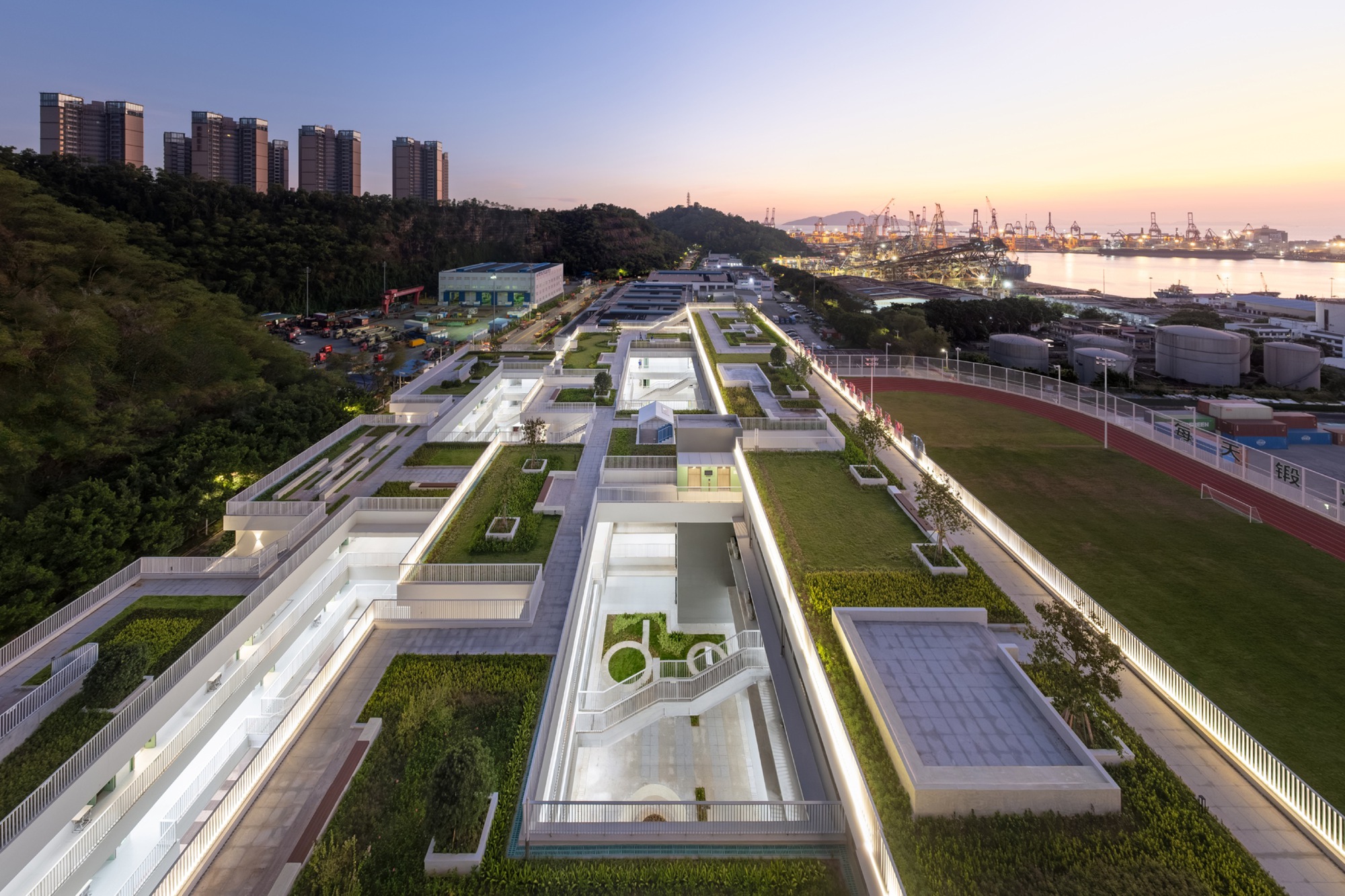 Dusk aerial view of the lit rooftop gardens, stairs and running track beside a harbour with cranes glowing on the horizon
