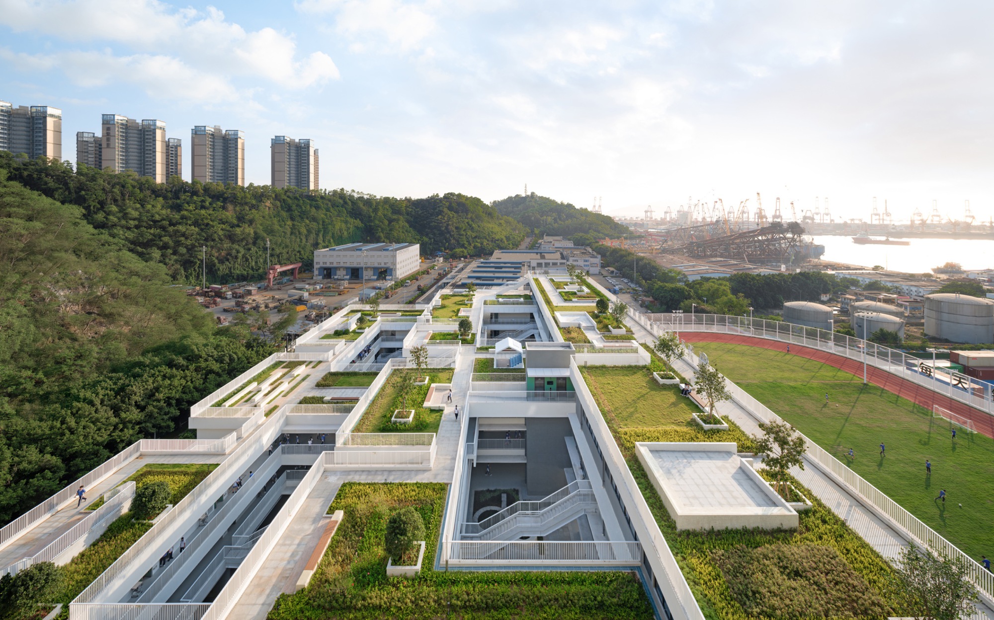 Elevated view of stepped green roof terraces, white stair voids and a red running track under a hazy sky