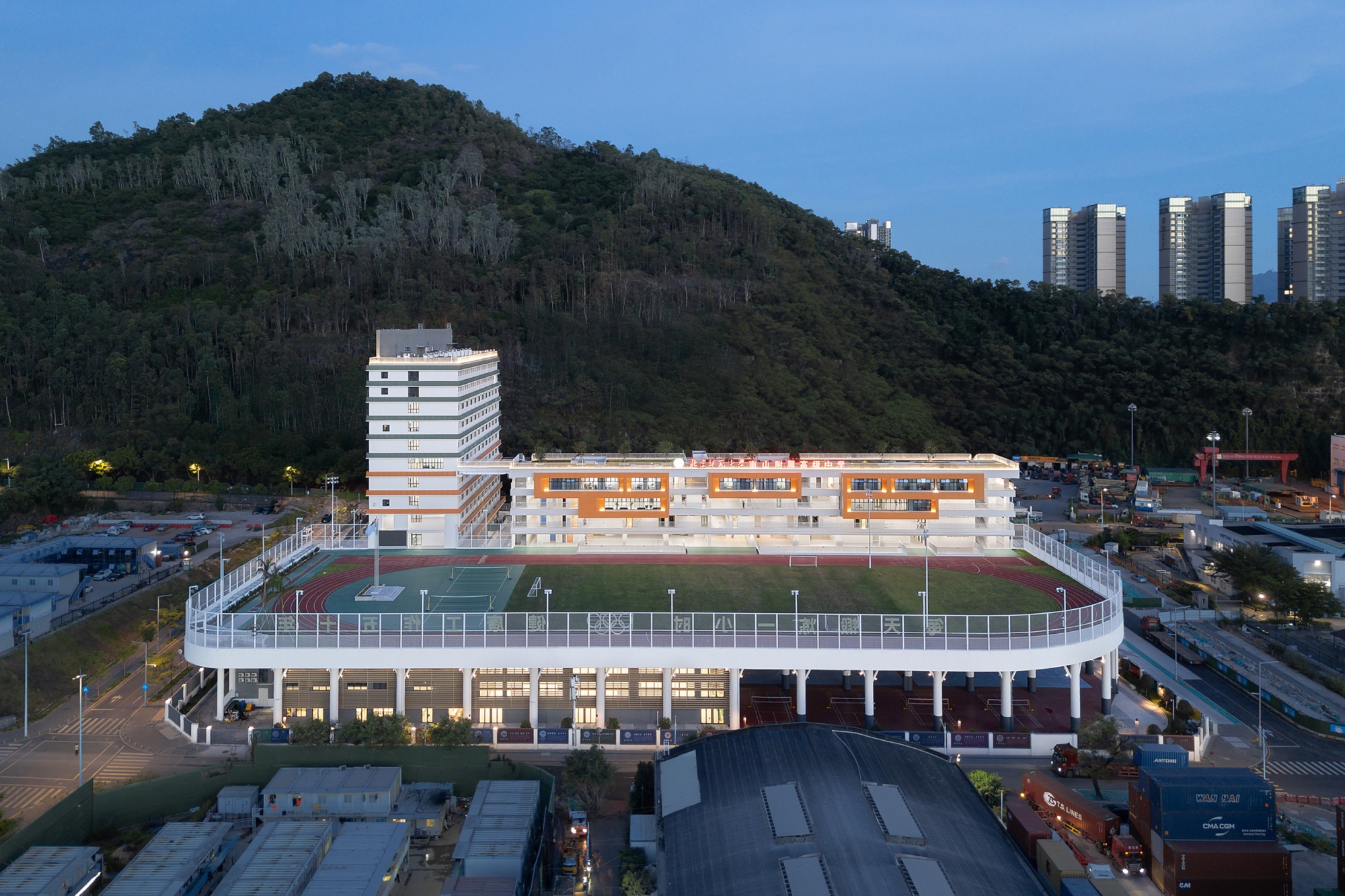 Dusk view of the school's raised sports field ringed by a white railing, with the hill rising behind