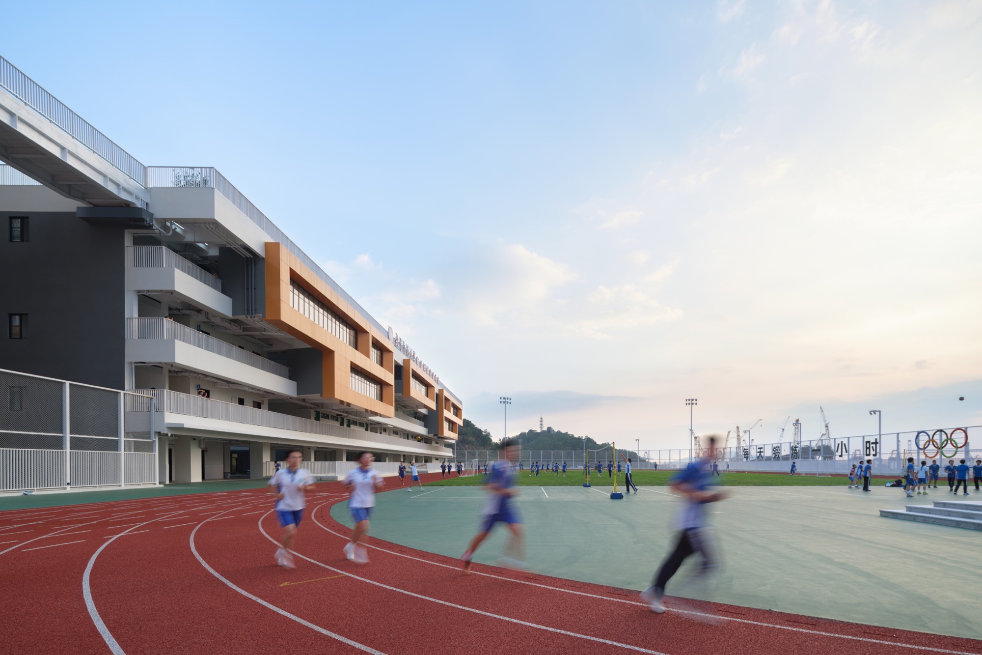 Runners blurred in motion on the red track beside a stepped school facade with orange cladding at dusk