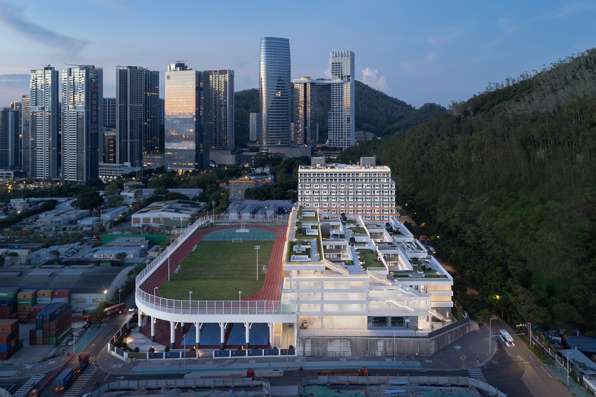 Dusk view of the track raised on slender columns beside terraced green roofs, with towers and forested hillside behind