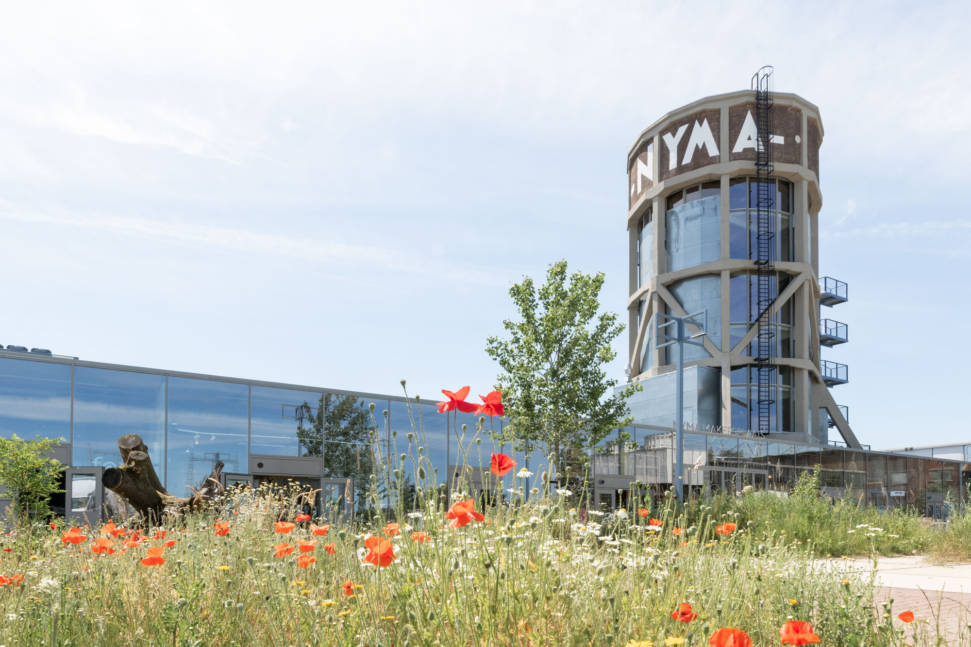 Cylindrical brick water tower with lettering and external ladder rising behind a glass-clad hall and a foreground meadow of red poppies