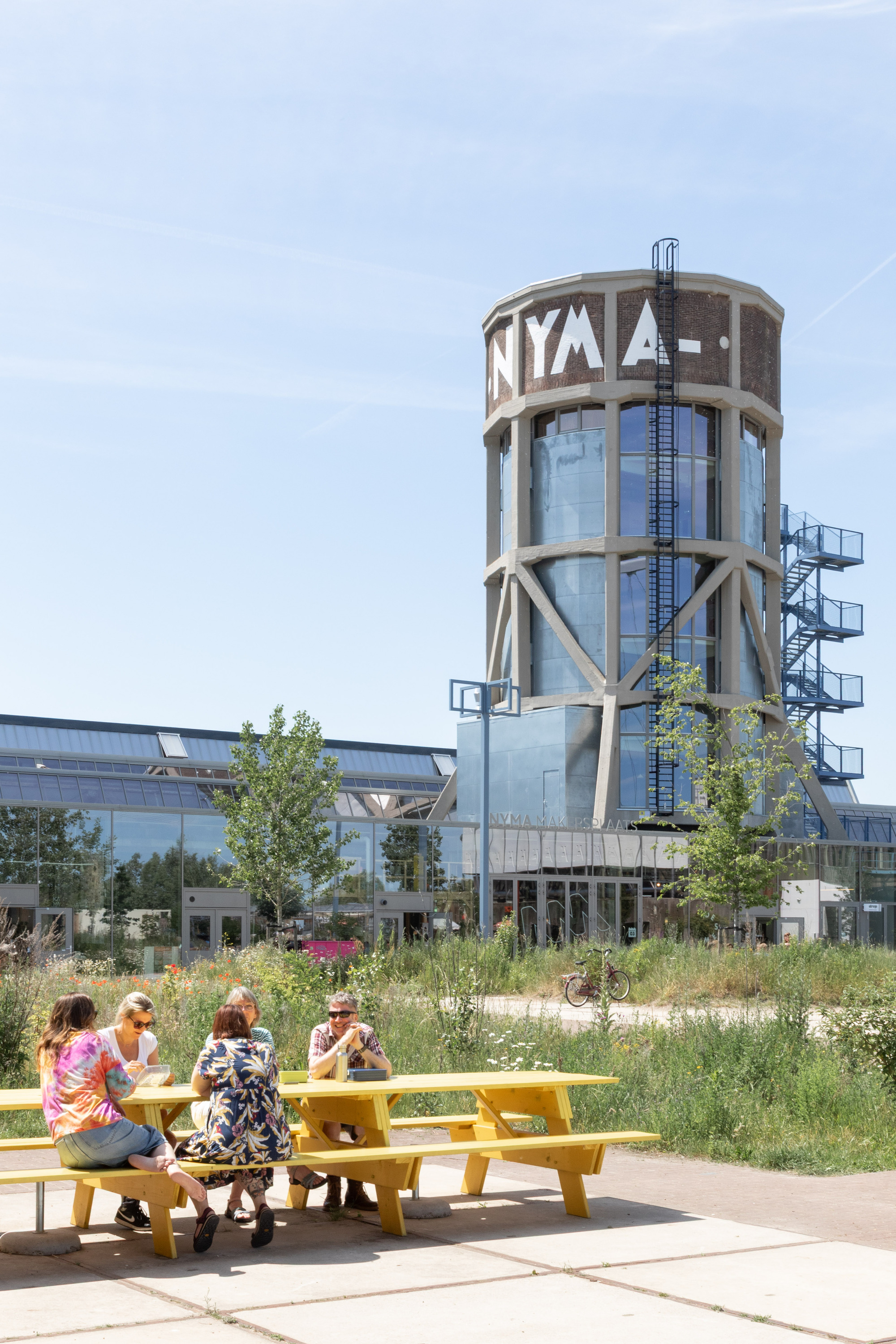 Cylindrical brick tower with lettering and steel bracing rising behind a yellow picnic table where people sit on a sunny day