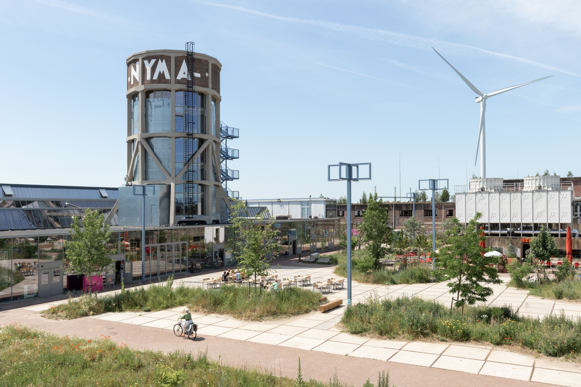 Wide view of the lettered tower, glazed hall and paved square with planted beds, a cyclist and a wind turbine under blue sky