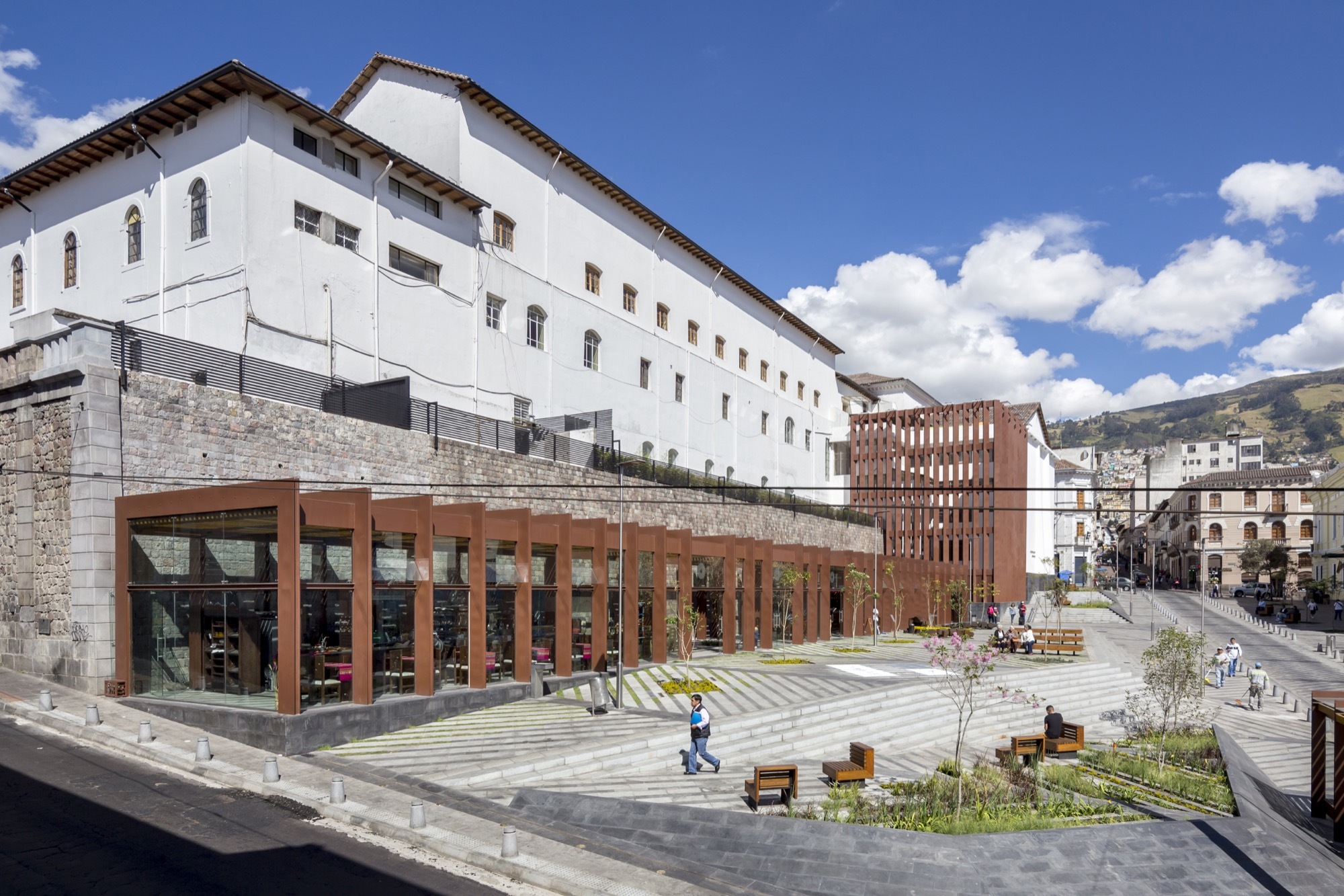 Terraced plaza with striped stone steps below a white convent wall and a rust-colored glass-fronted library pavilion under blue sky
