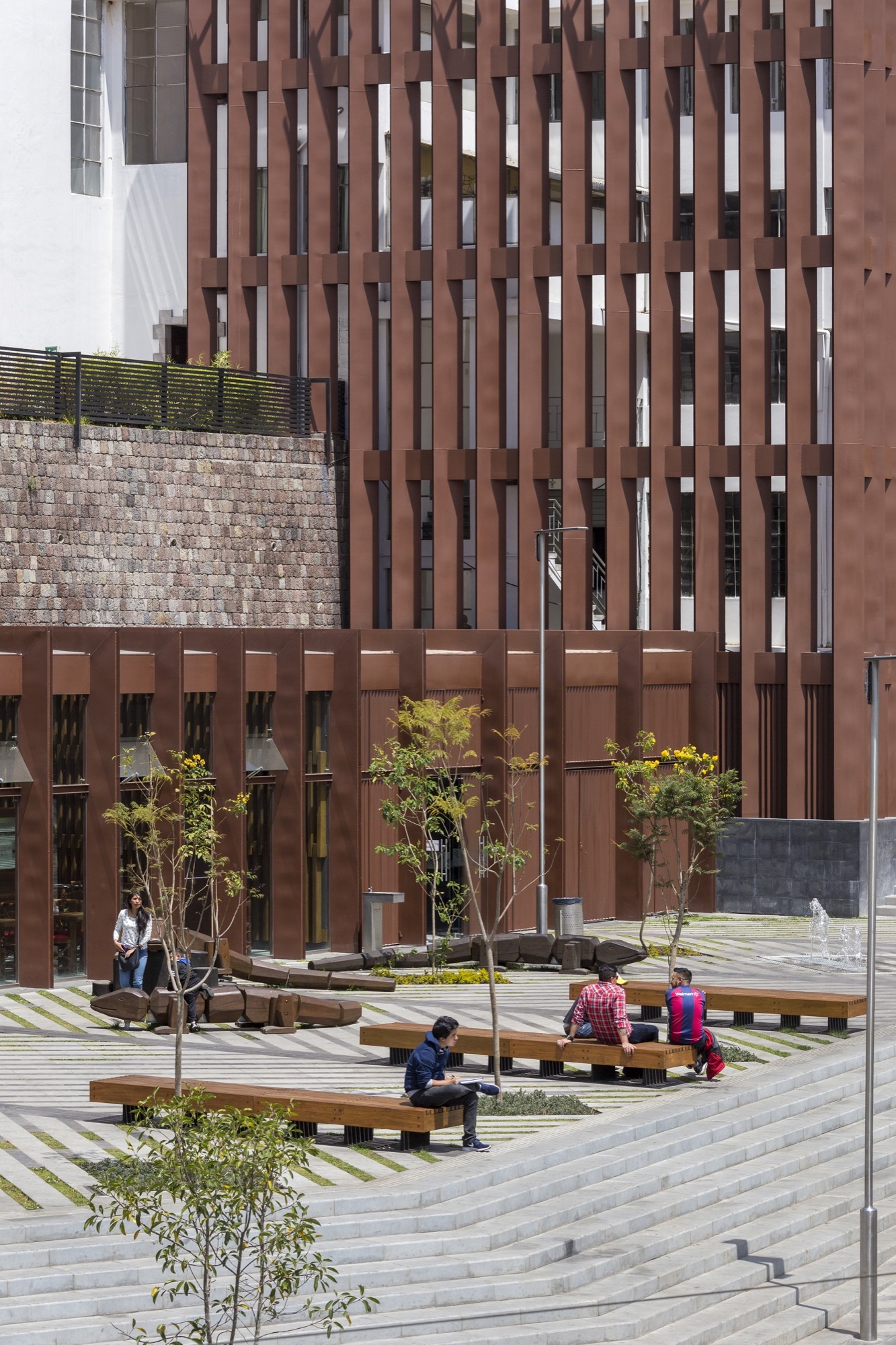 Rust-toned vertical slat tower above a striped paved plaza where people sit on timber benches among young trees