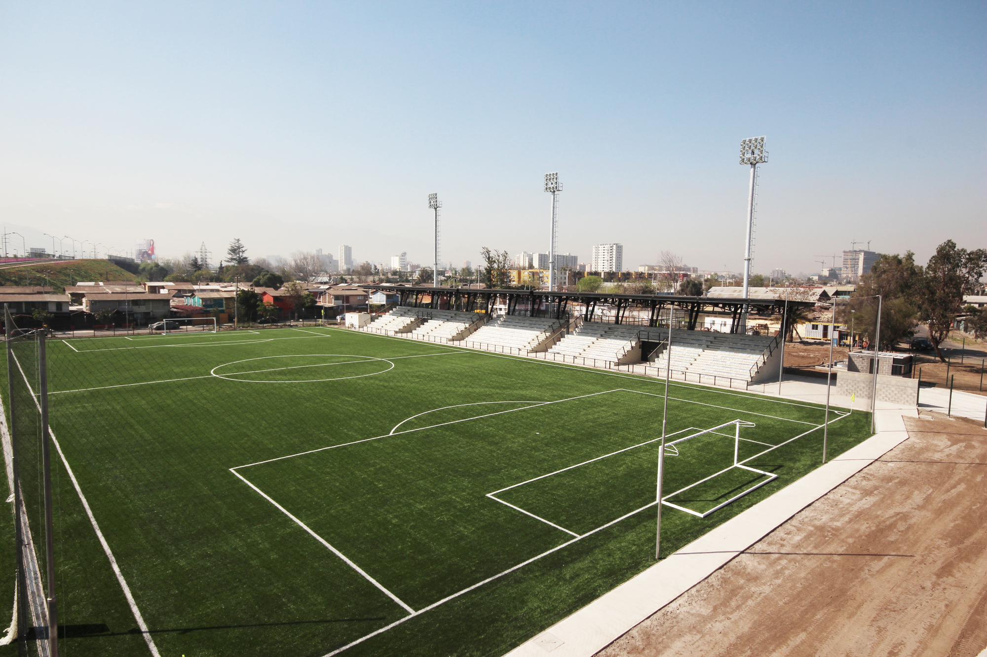 Elevated view of the artificial turf pitch and steel-roofed stands, with floodlight towers and a hazy city skyline behind