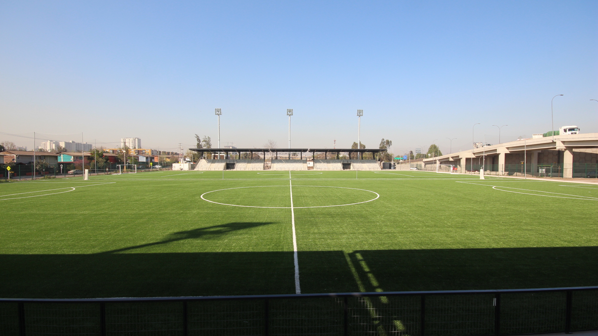 Wide view across the pitch in late sun, with the roofed grandstand at the far end and a concrete viaduct on the right