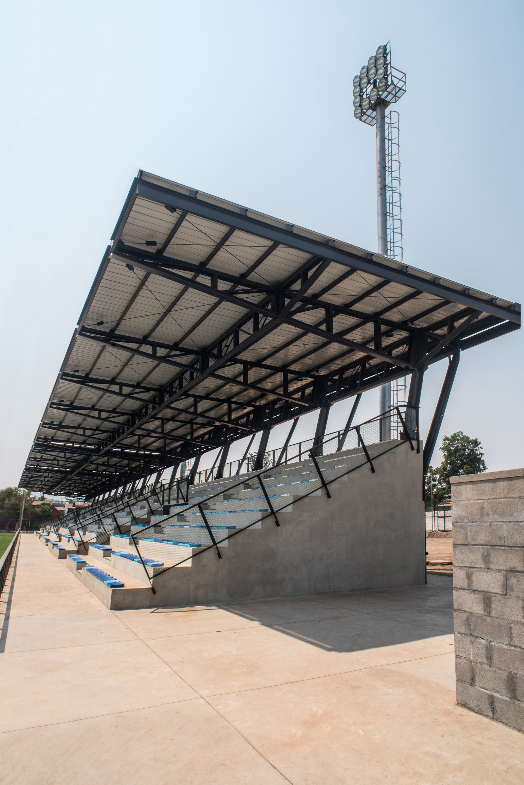 Side view of the cantilevered steel roof over concrete stepped stands with blue seats, beside a floodlight tower and a concrete block wall