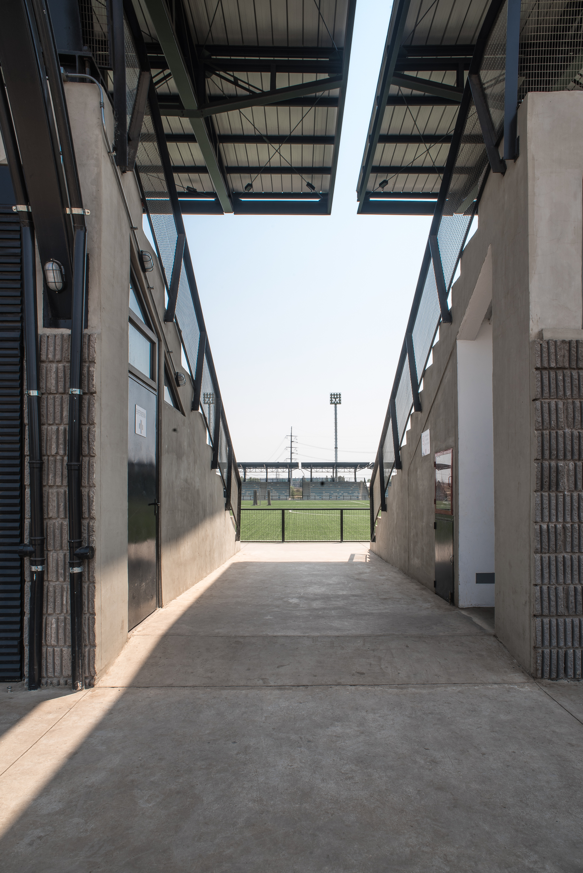 Concrete passage between stand volumes under angled steel roof canopies, framing the pitch and a distant floodlight in hazy daylight