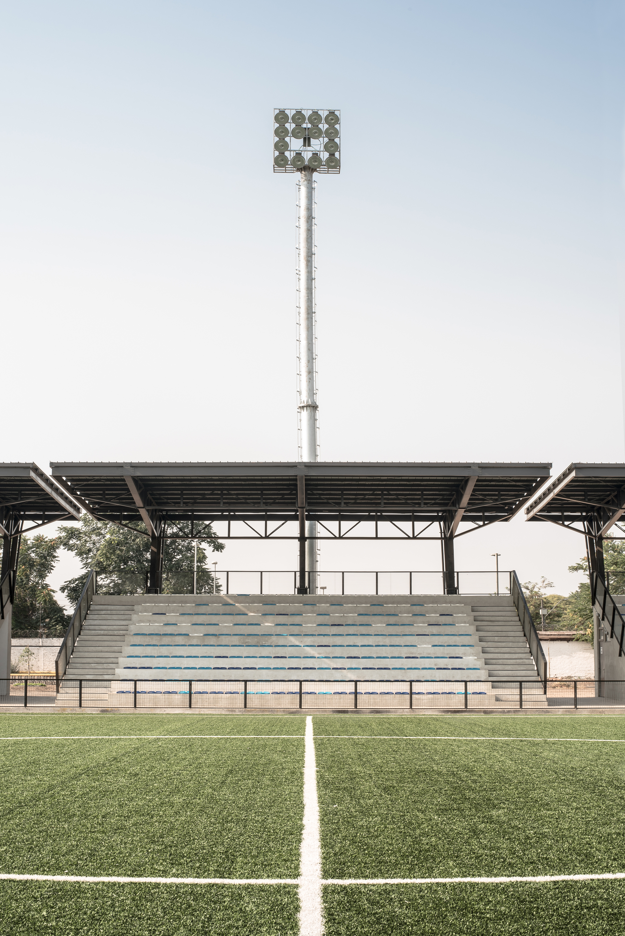 Front view of the stand with a steel canopy, central concrete steps and a tall floodlight mast rising behind it in pale haze