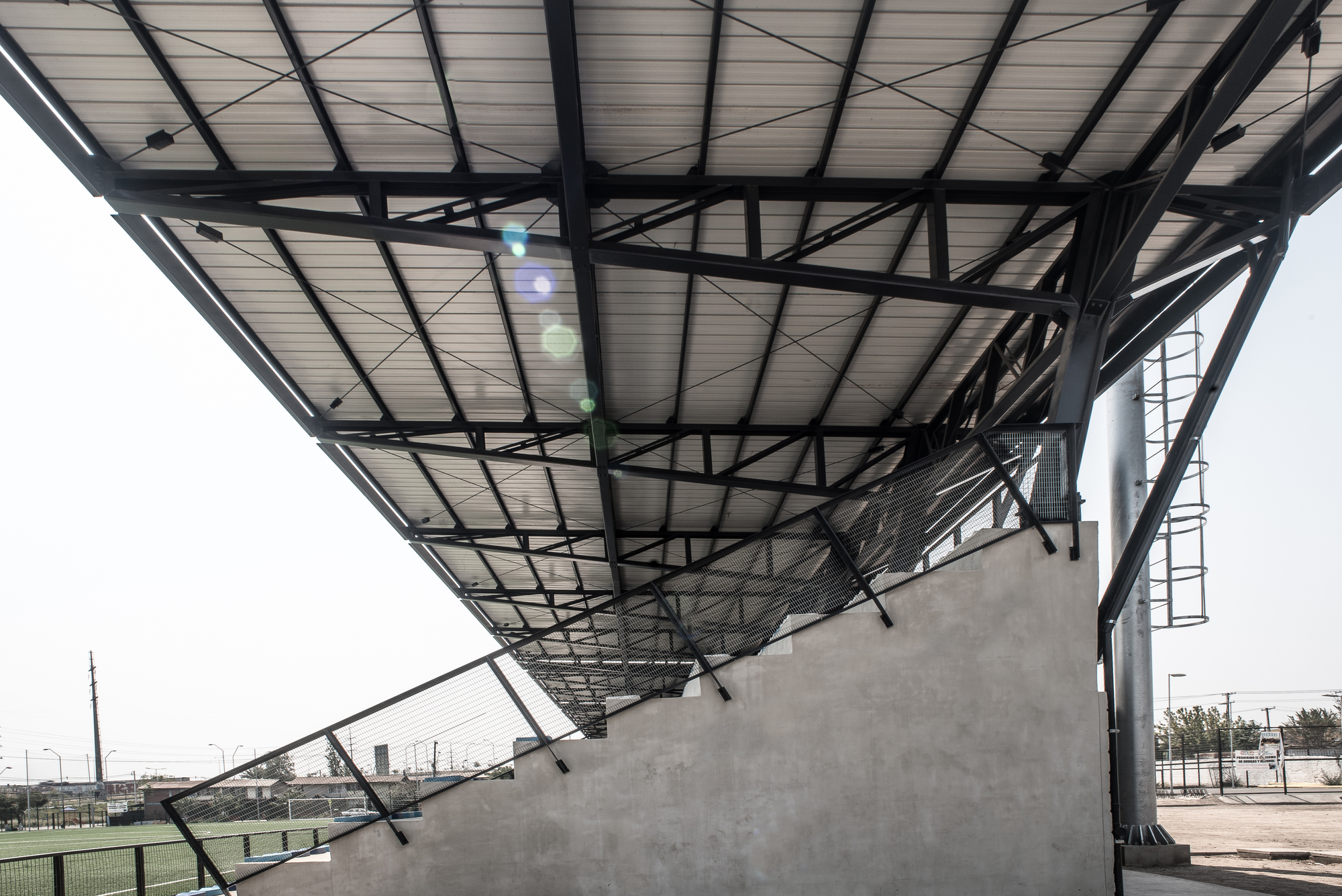 Underside of the cantilevered steel truss roof above a concrete stepped grandstand, with lens flare and a haze-white sky