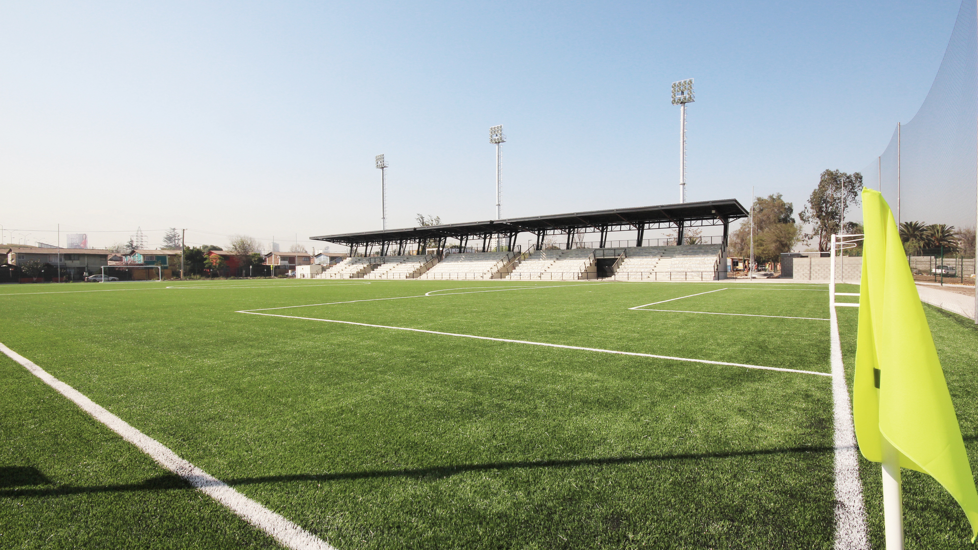 Pitch-level view toward a covered grandstand and floodlight masts across green turf, with a yellow corner flag in a clear sky