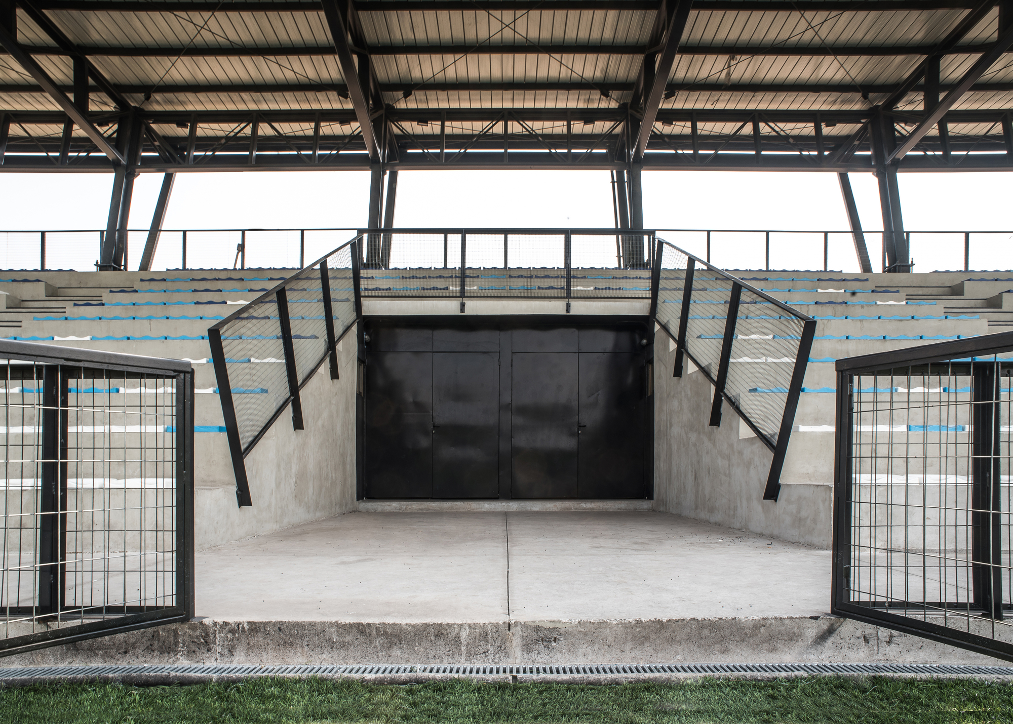 Black steel double doors set into the concrete stand, flanked by mesh railings and blue-striped seating rows under the roof