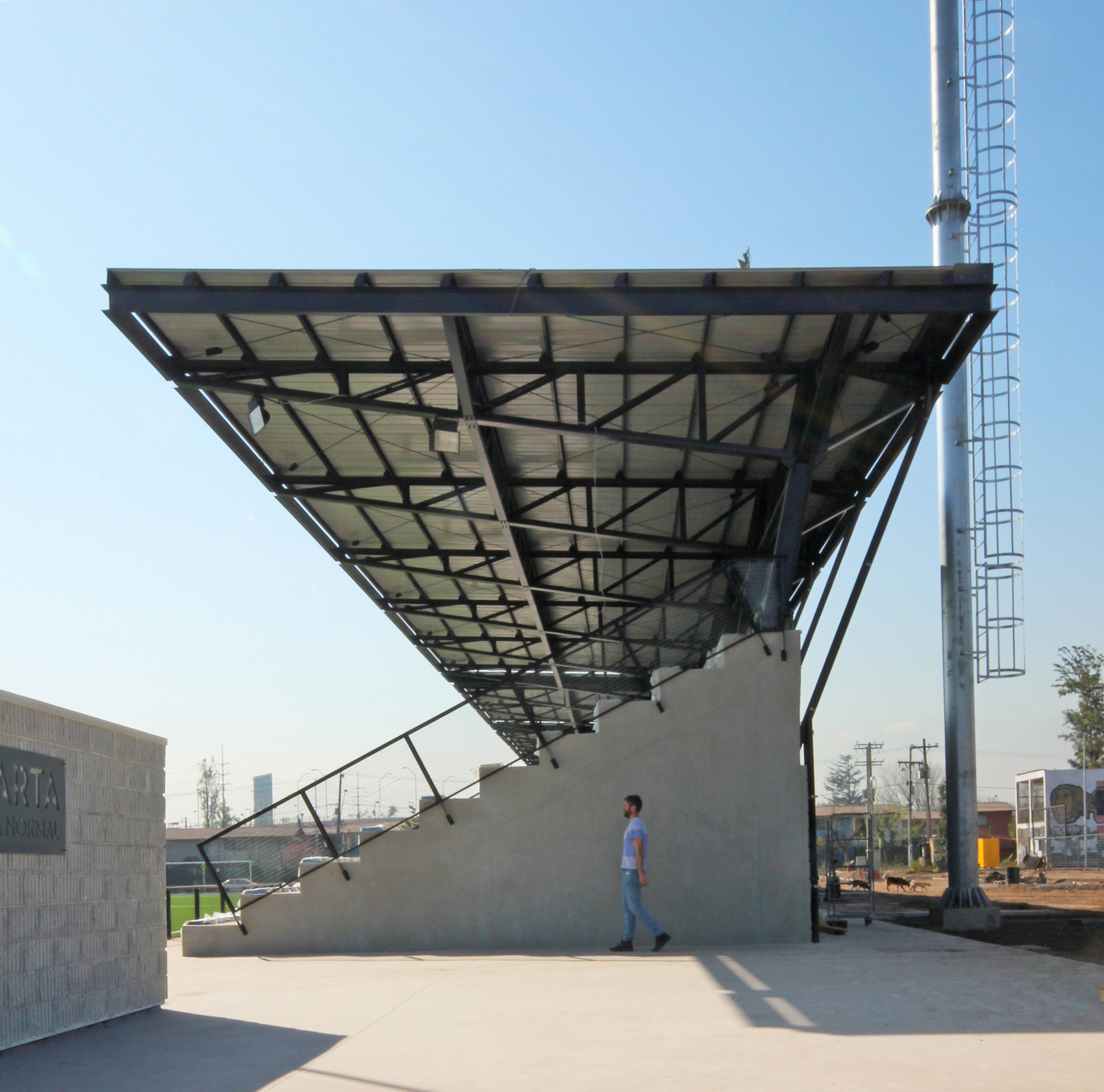 Side elevation of the cantilevered roof over the concrete stand, with a man walking past in strong afternoon sun