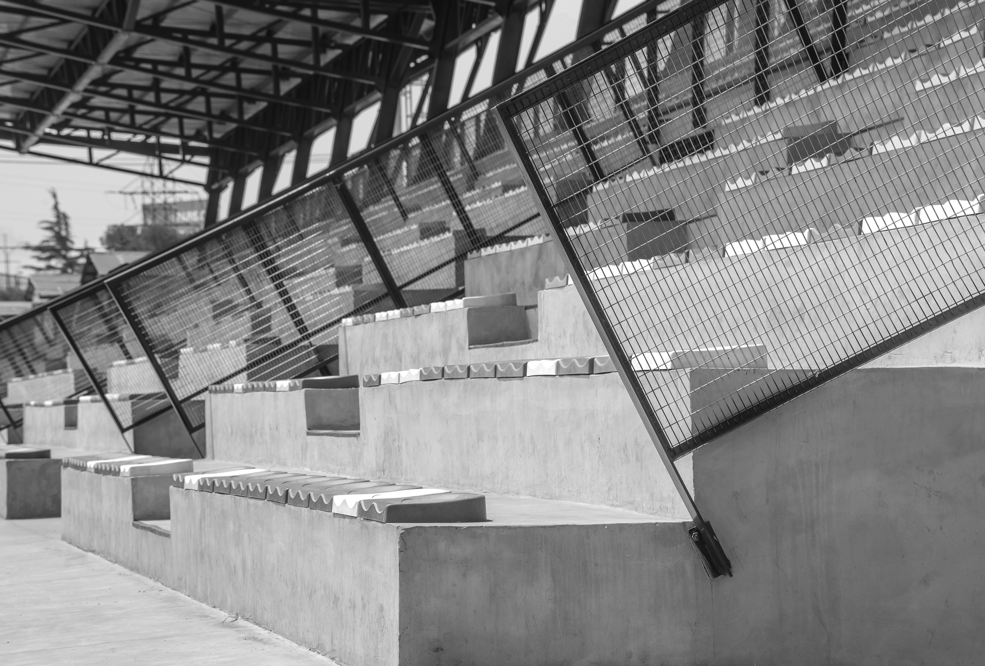 Black and white close-up of stepped concrete seating tiers behind an angled steel mesh guardrail, with the roof truss overhead