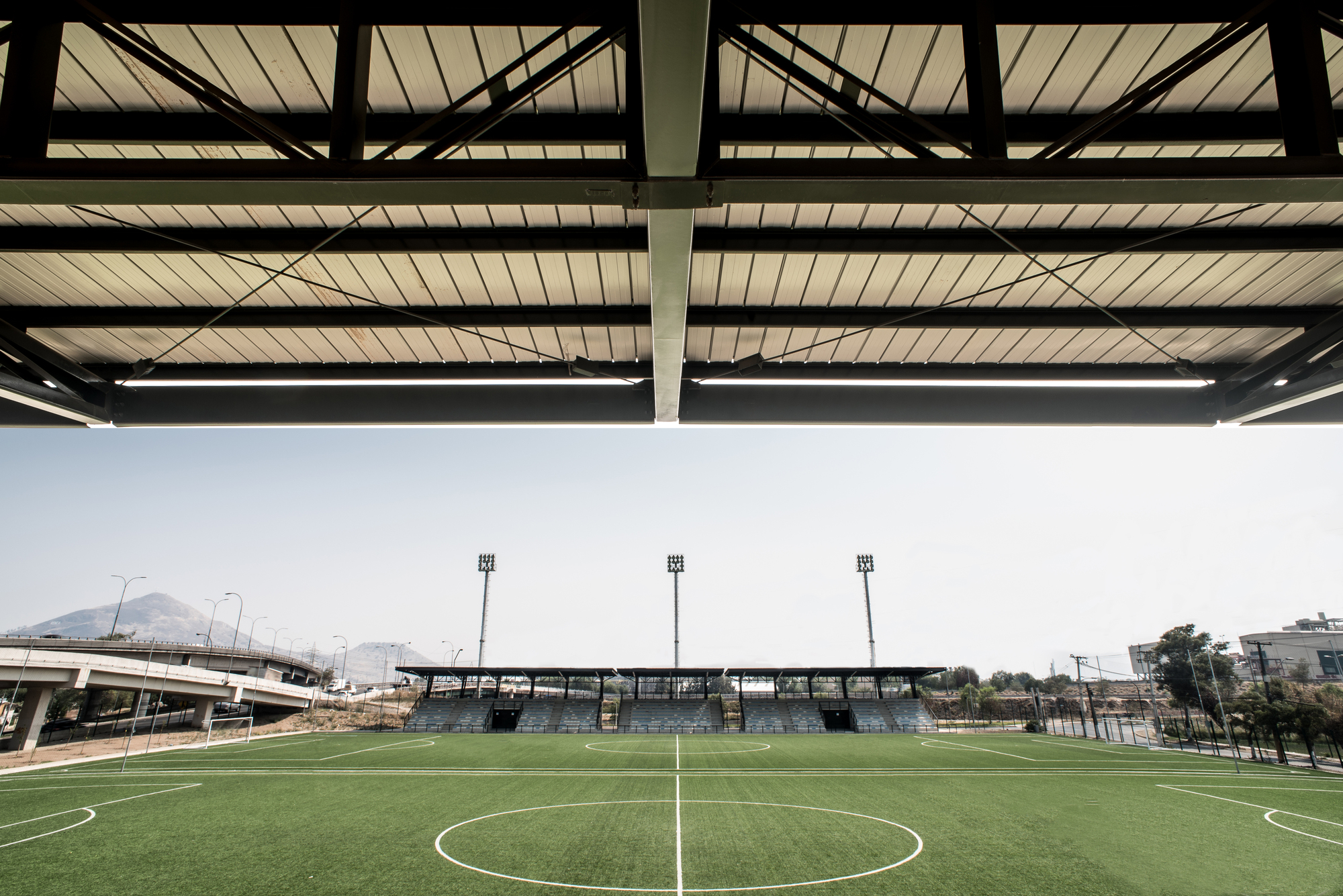 View from beneath the grandstand roof across the pitch to the opposite stand, with distant hills and an overpass in haze