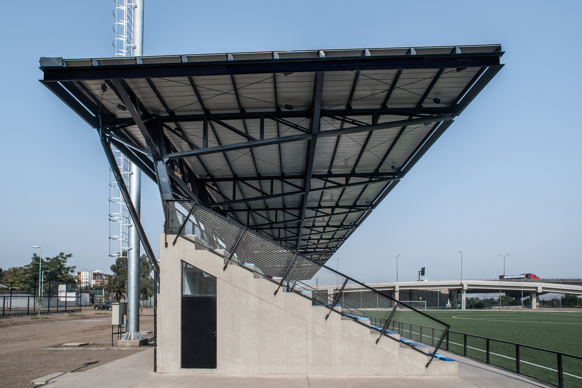 Underside of the cantilevered steel canopy over a concrete grandstand end, with a floodlight mast and elevated road beyond under clear sky