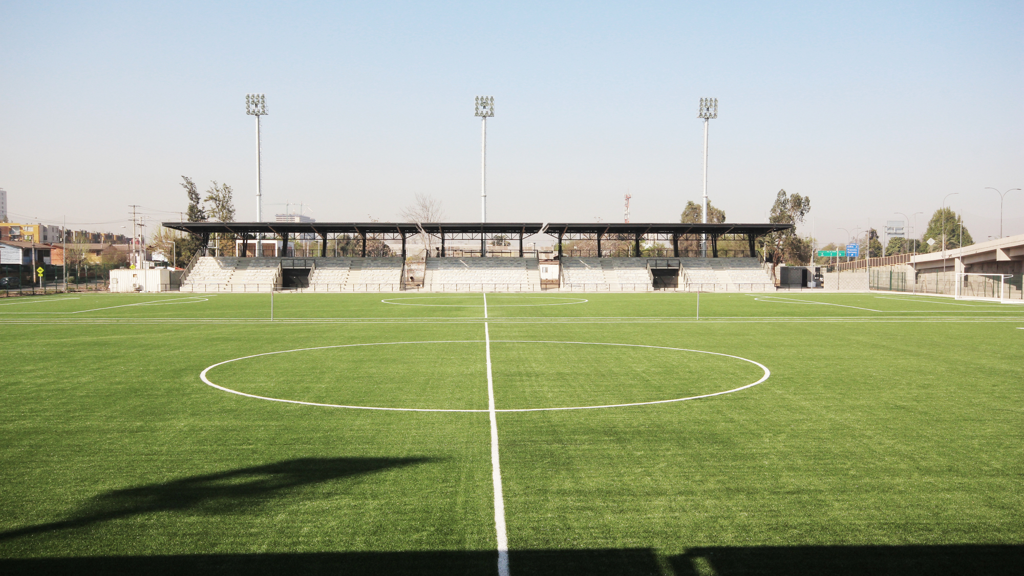 Centre circle of a synthetic turf pitch facing a low grandstand with three floodlight masts in hard midday sun