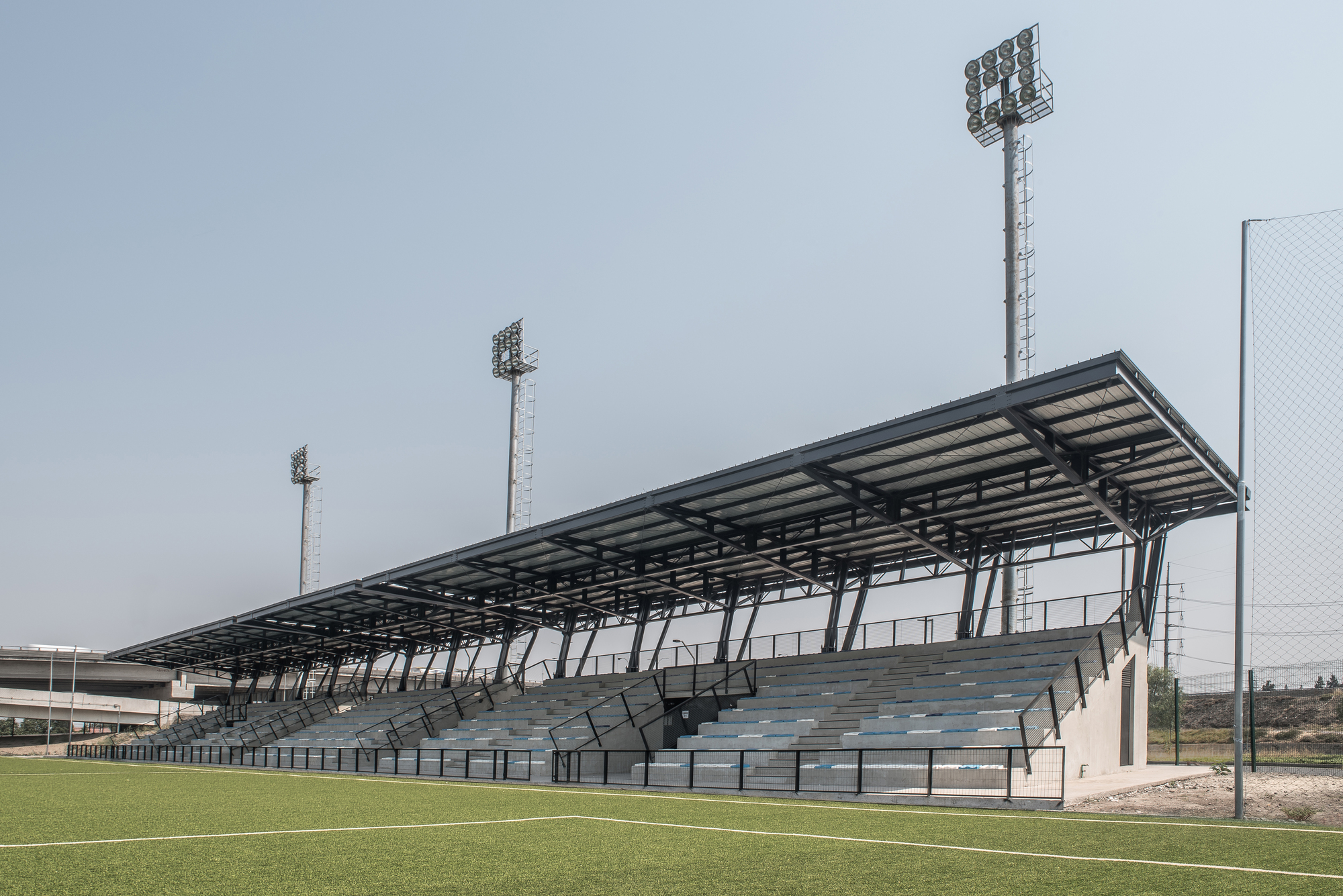 Angled view of the long steel-roofed grandstand with concrete bleachers, railings and tall floodlights above the green pitch in haze