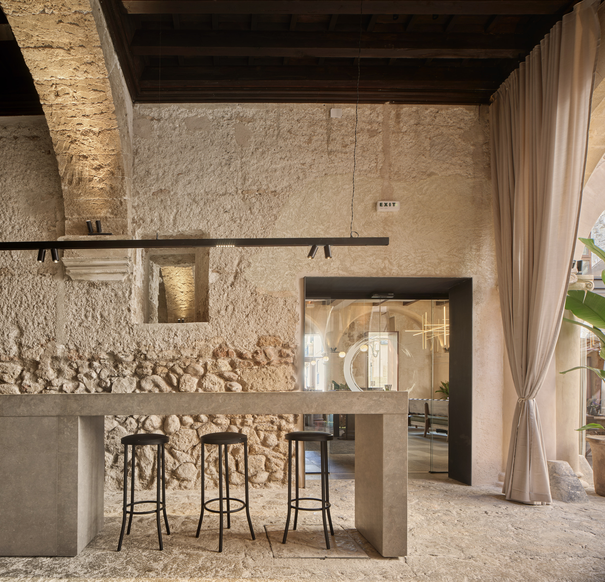 Stone bar counter with three black stools against a rough limestone wall, a linear pendant light overhead and a linen curtain beside