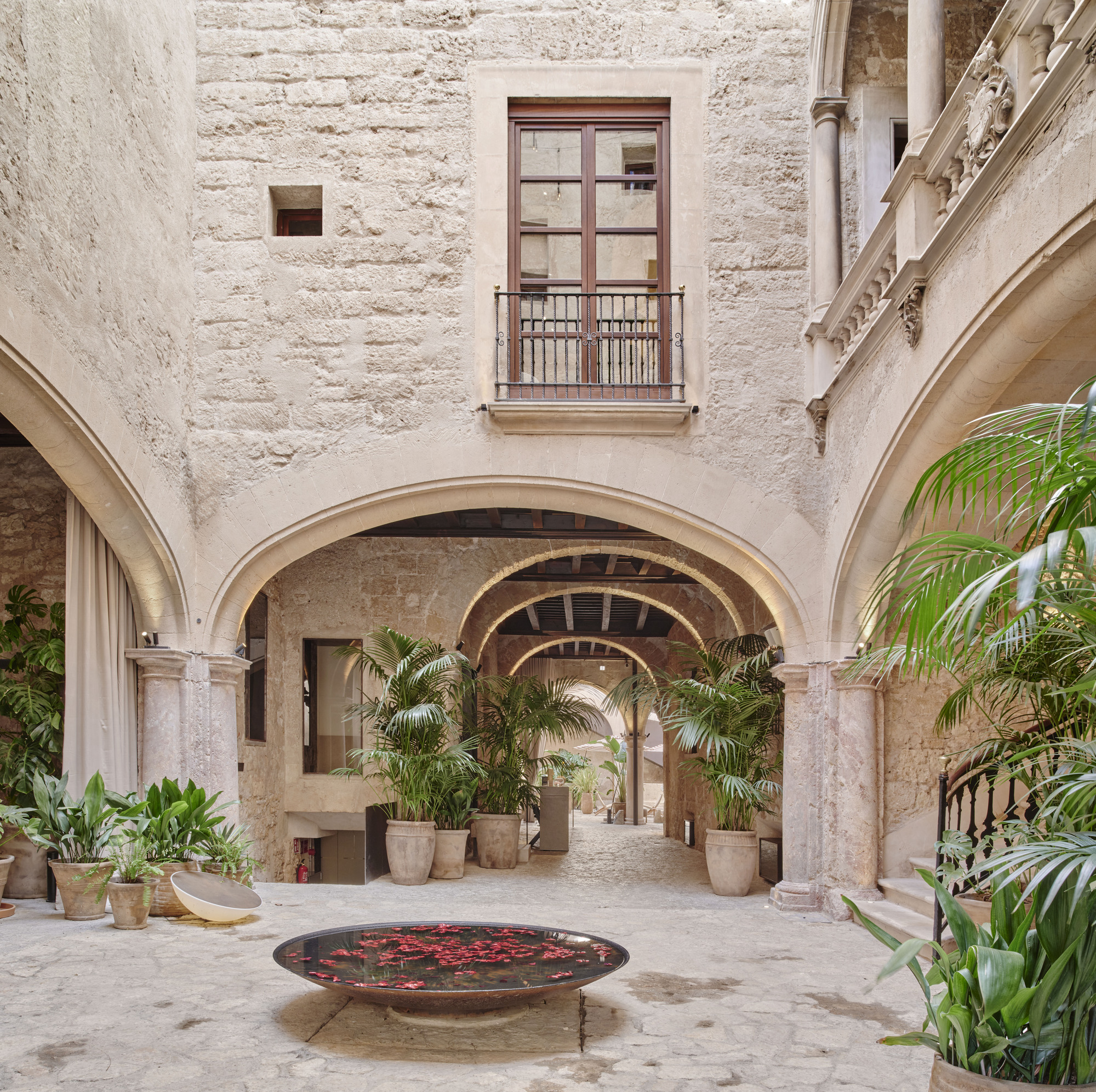 Courtyard with pointed stone arches, potted palms and a shallow bowl of red petals on the paving in soft daylight