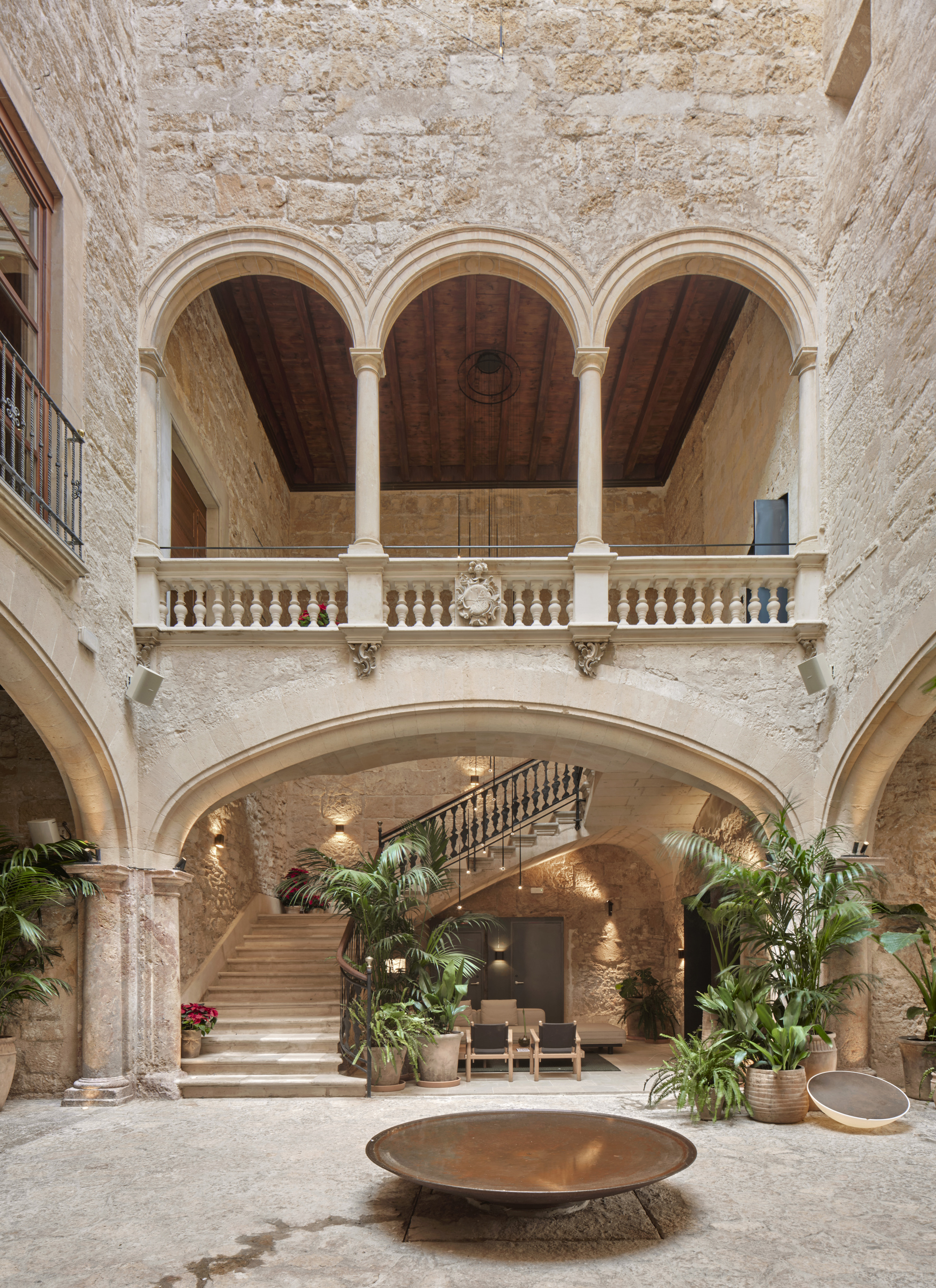 Courtyard view toward a triple-arched loggia and stone staircase, with a rust-toned bowl on the floor and potted plants beneath