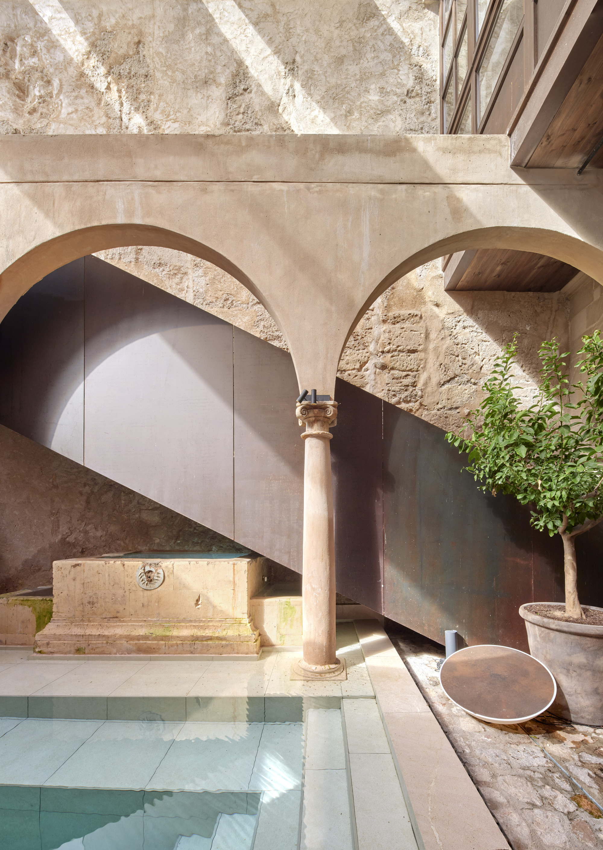 Slim stone column between two arches above a plunge pool, with a dark steel panel wall and a potted lemon tree in sunlight