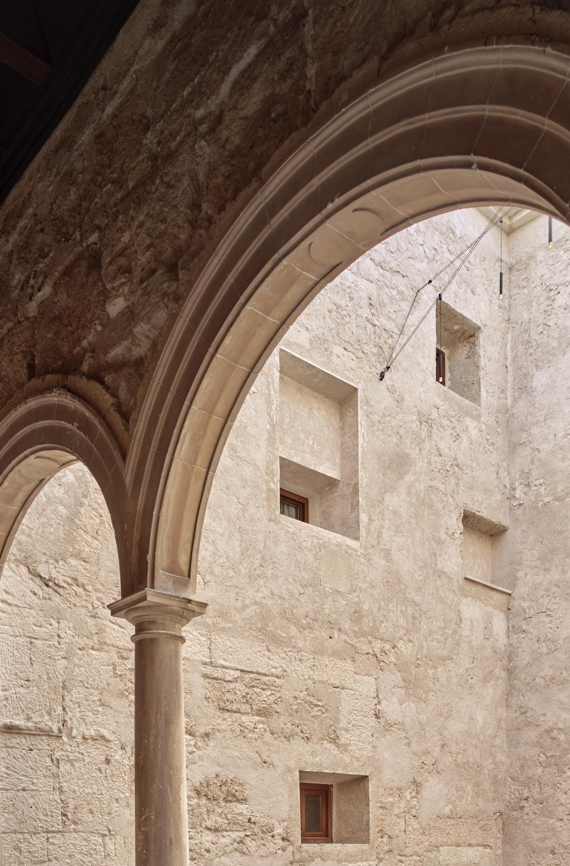 Looking through a moulded stone arch and slender column at a rough plastered courtyard wall with small recessed windows