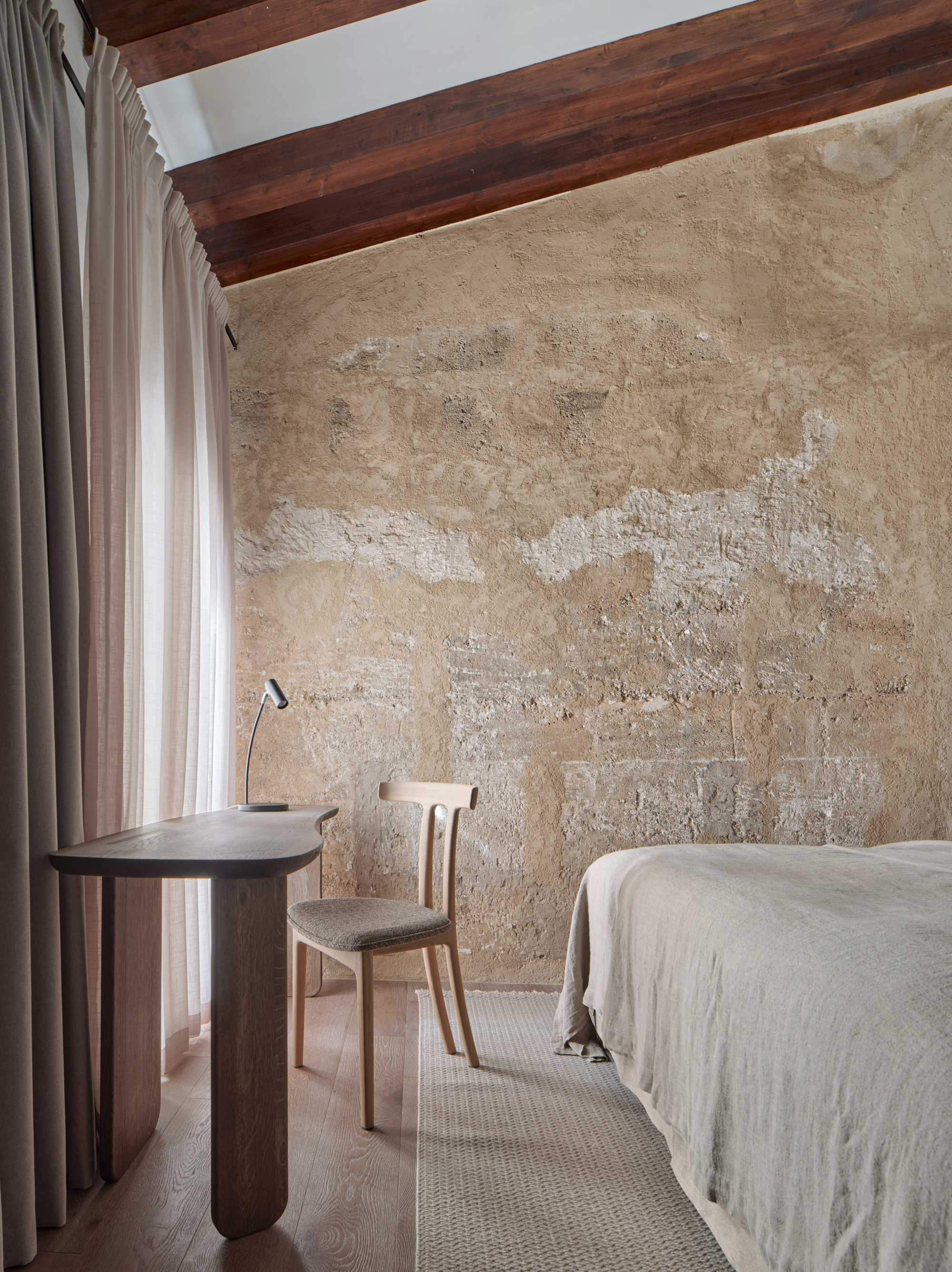 Bedroom corner with a weathered plaster wall, timber beams, a curved wooden desk, oak chair and linen bedding beside sheer curtains