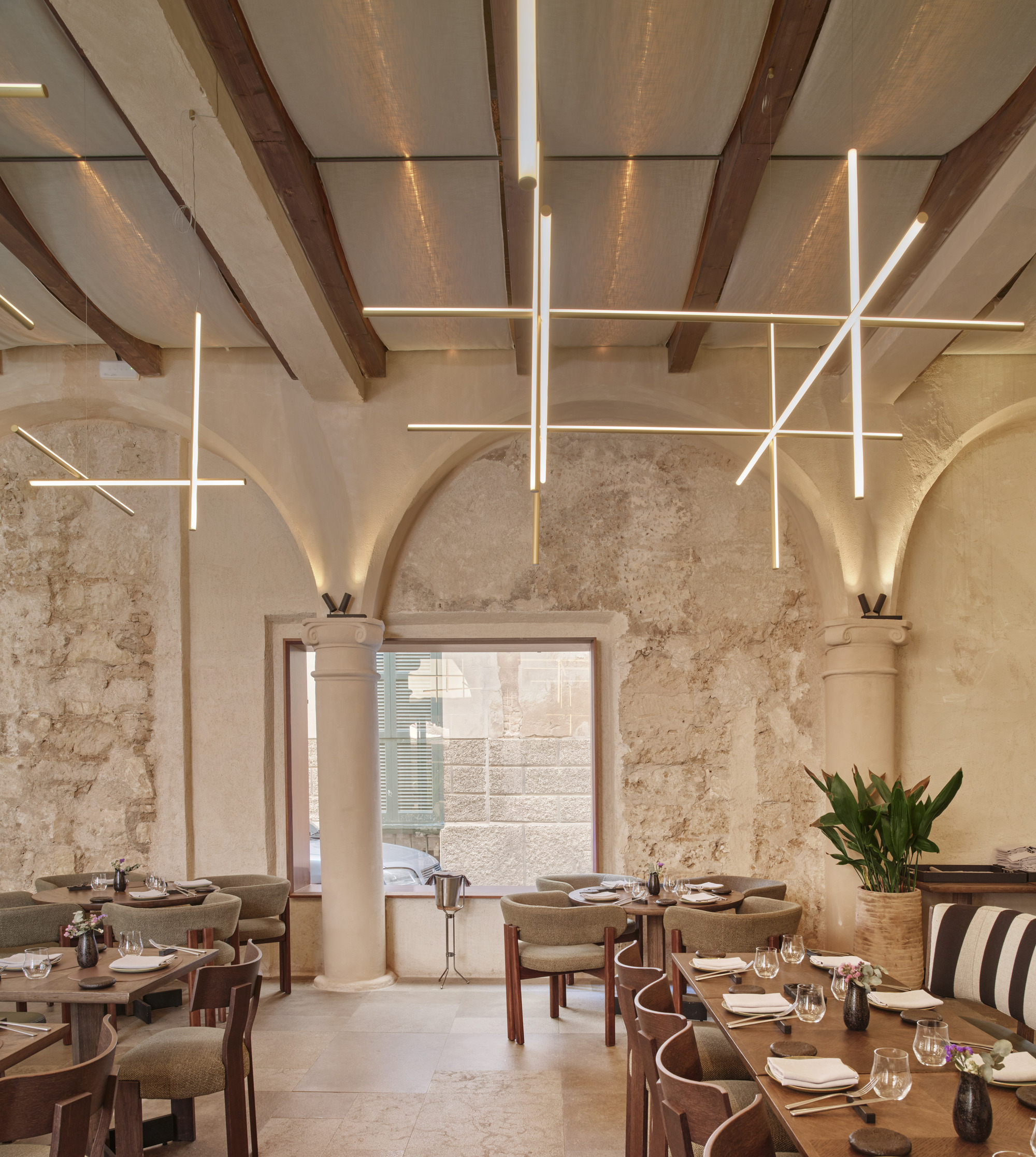 Restaurant dining room with vaulted stone arches, timber beams and linear light fixtures hanging above wooden tables and upholstered chairs