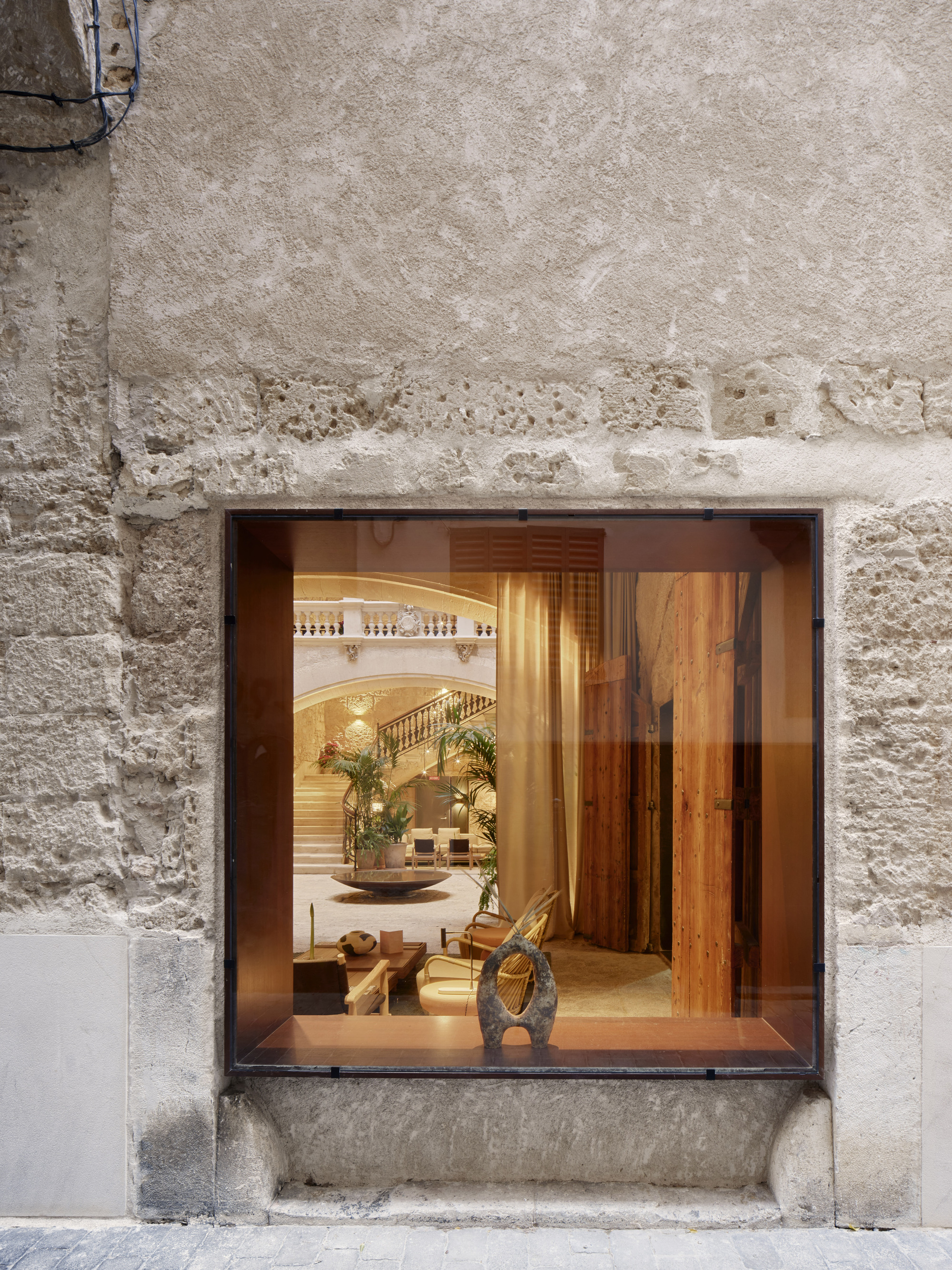 Street-level window set into a rough limestone wall, framing a view of a lobby with a staircase and potted palms
