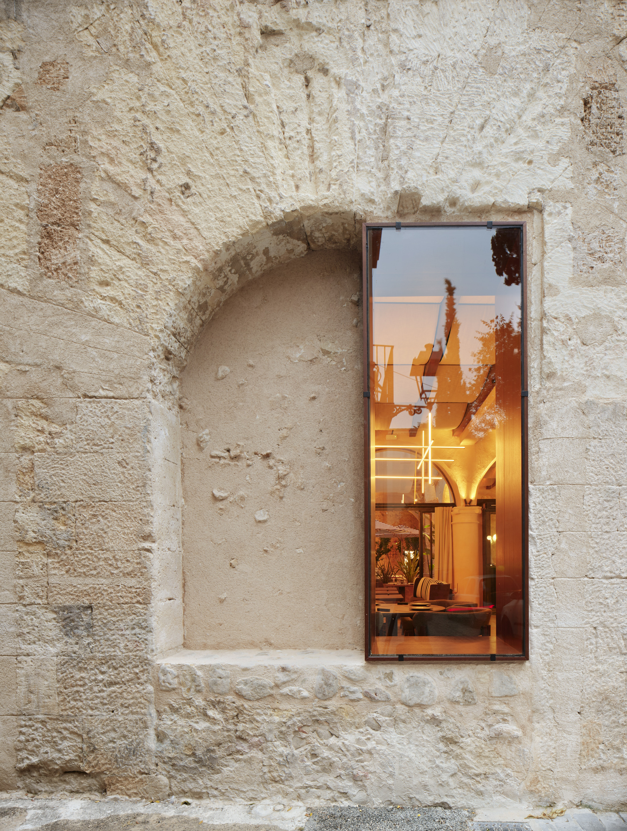 Tall narrow glazed opening beside a blocked arch in a weathered stone wall, reflecting warm interior light at dusk