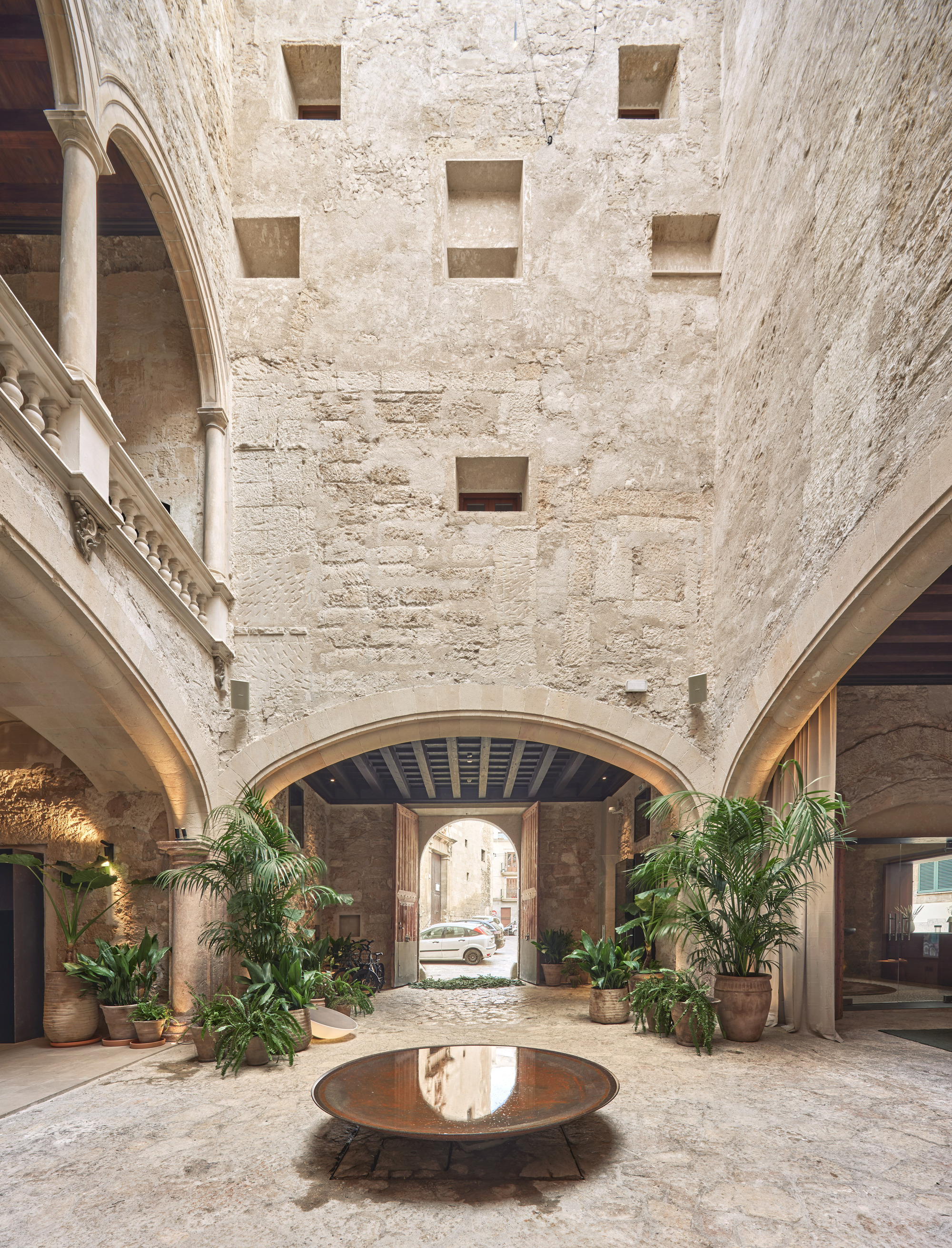 Stone courtyard with a round bronze basin at center, potted palms and a street visible through the arched entrance