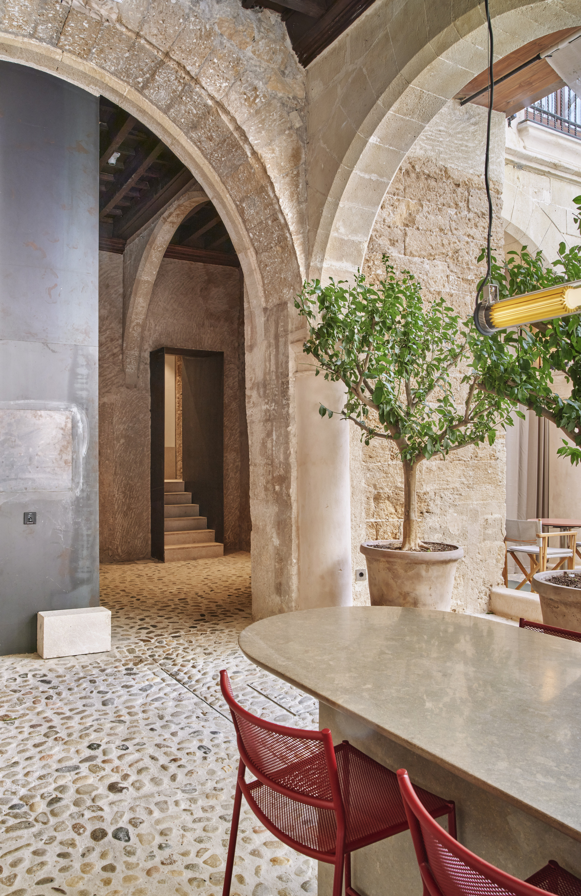 Potted lemon tree beside a pebble-paved floor and stone arches, with red mesh chairs at an oval table in soft daylight