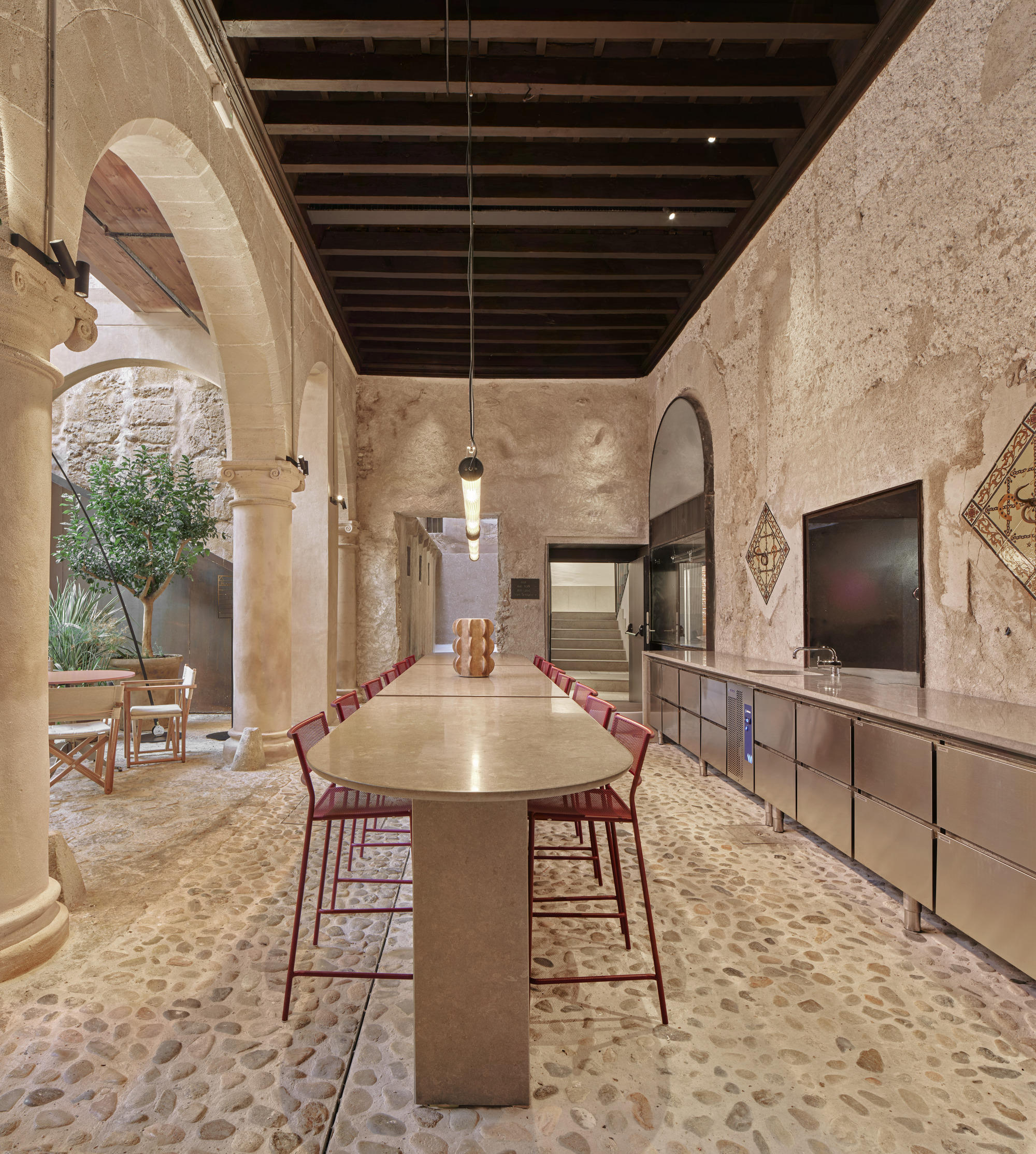 Long communal table with red stools under a dark timber ceiling, facing a stainless steel kitchen counter along a rough stone wall