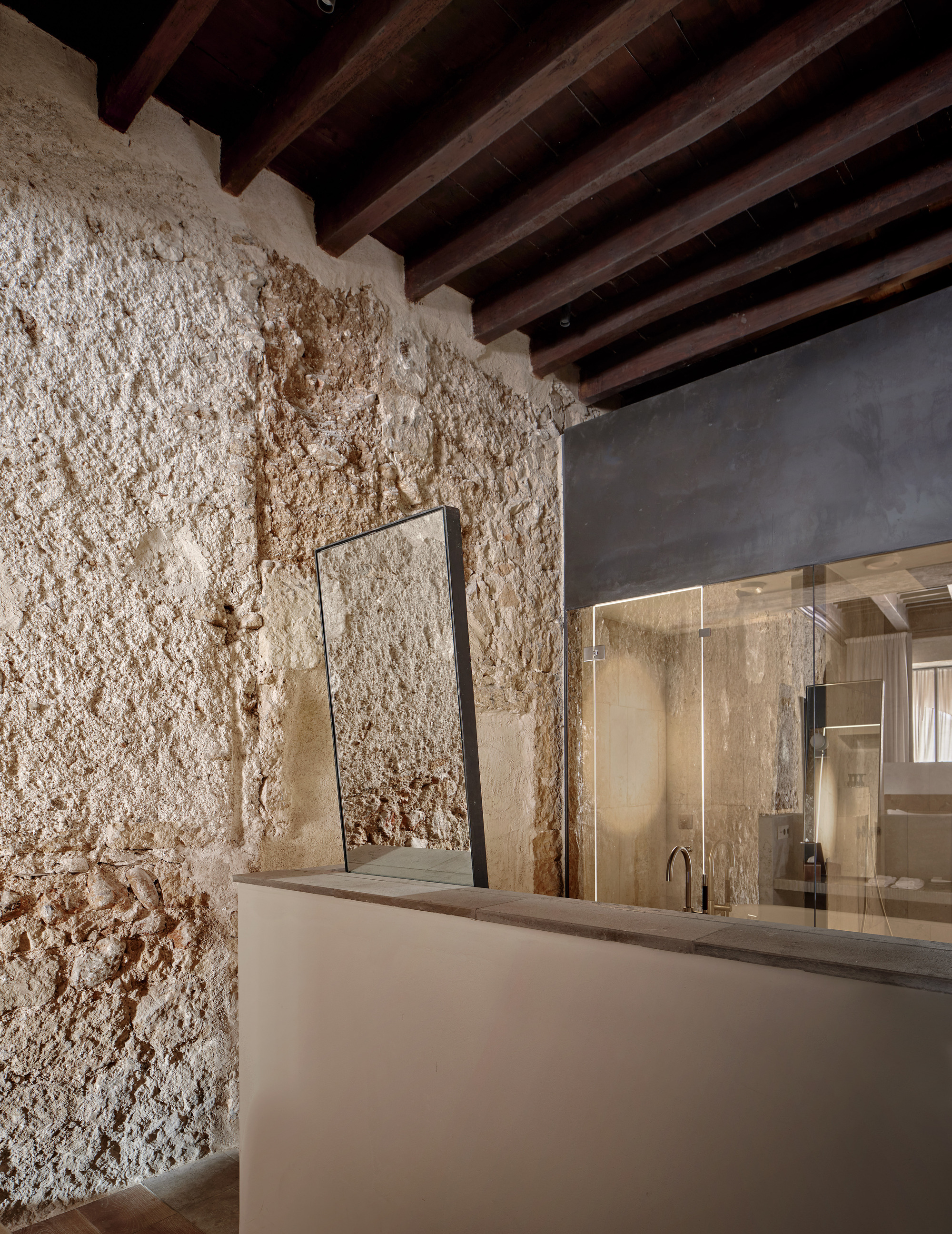 Rough lime-plastered stone wall with a leaning mirror beside a glass-enclosed bathroom under exposed dark timber beams