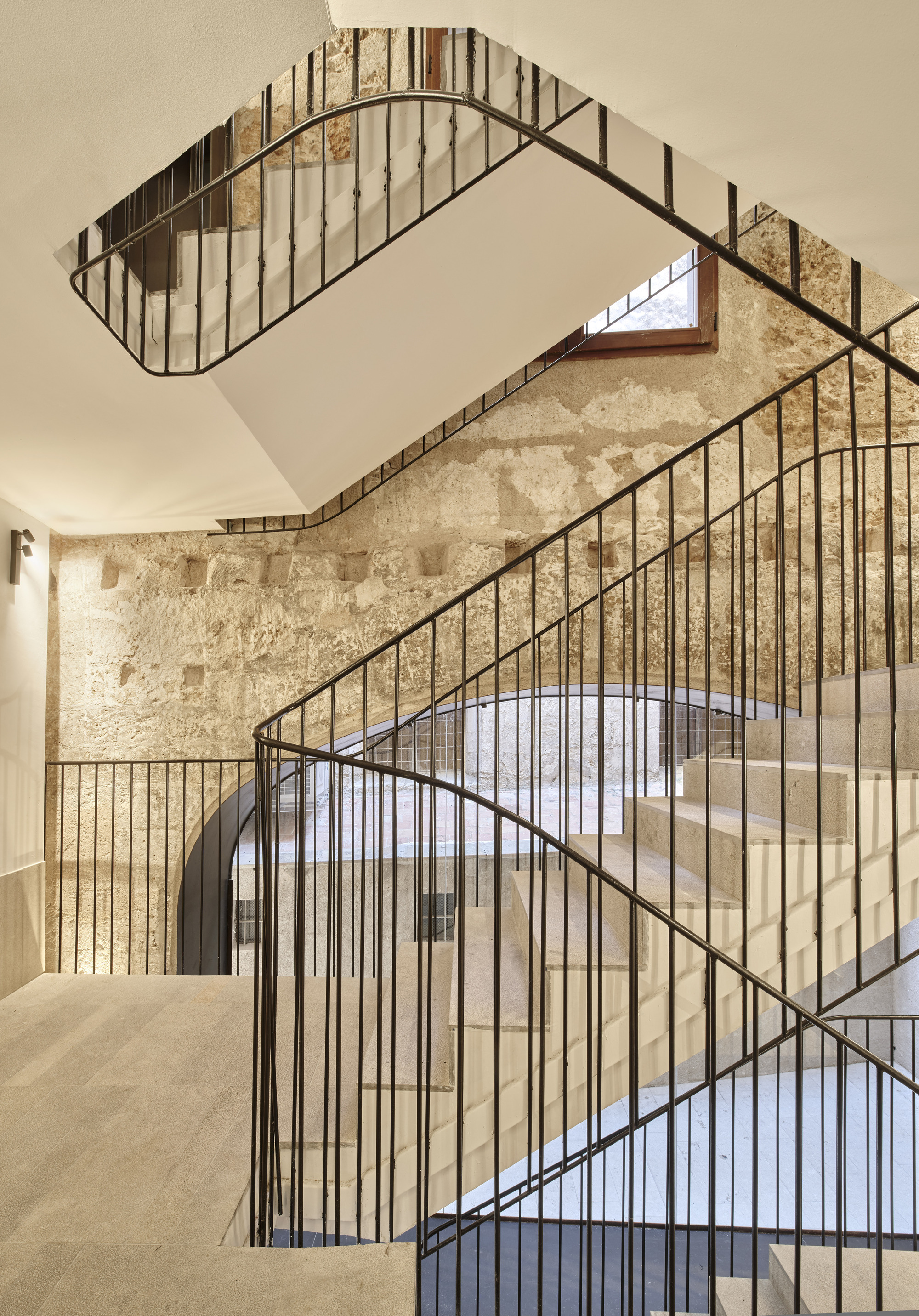 Stone staircase with slim black metal balustrade curving up past a worn stone wall and a small timber-framed window