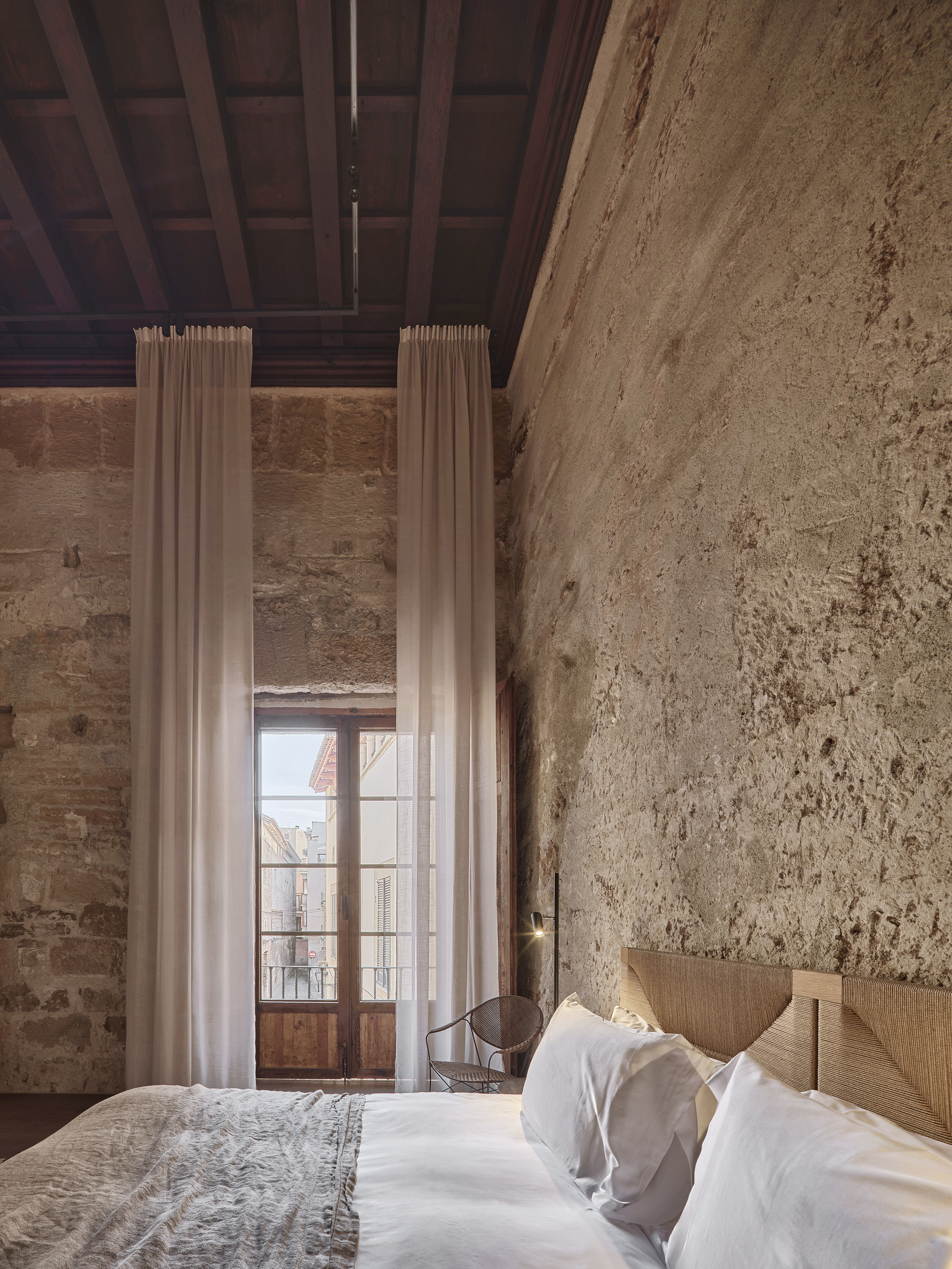 Bedroom with a timber headboard and tall sheer curtains framing a wooden French window, set against a weathered stone wall in daylight
