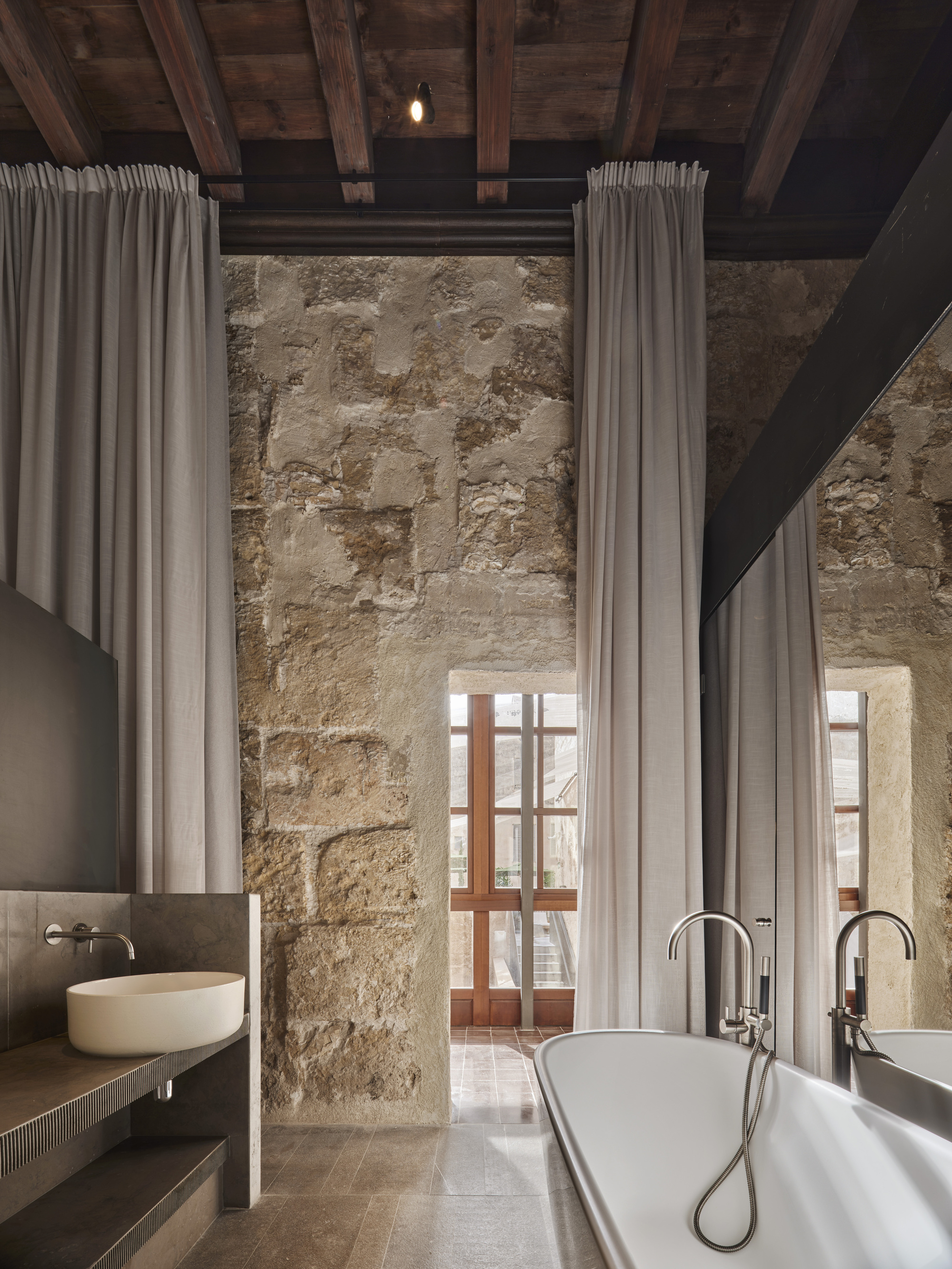 Bathroom with rough sandstone wall, floor-to-ceiling grey linen curtains, freestanding tub and round basin, daylight coming through a timber-framed window