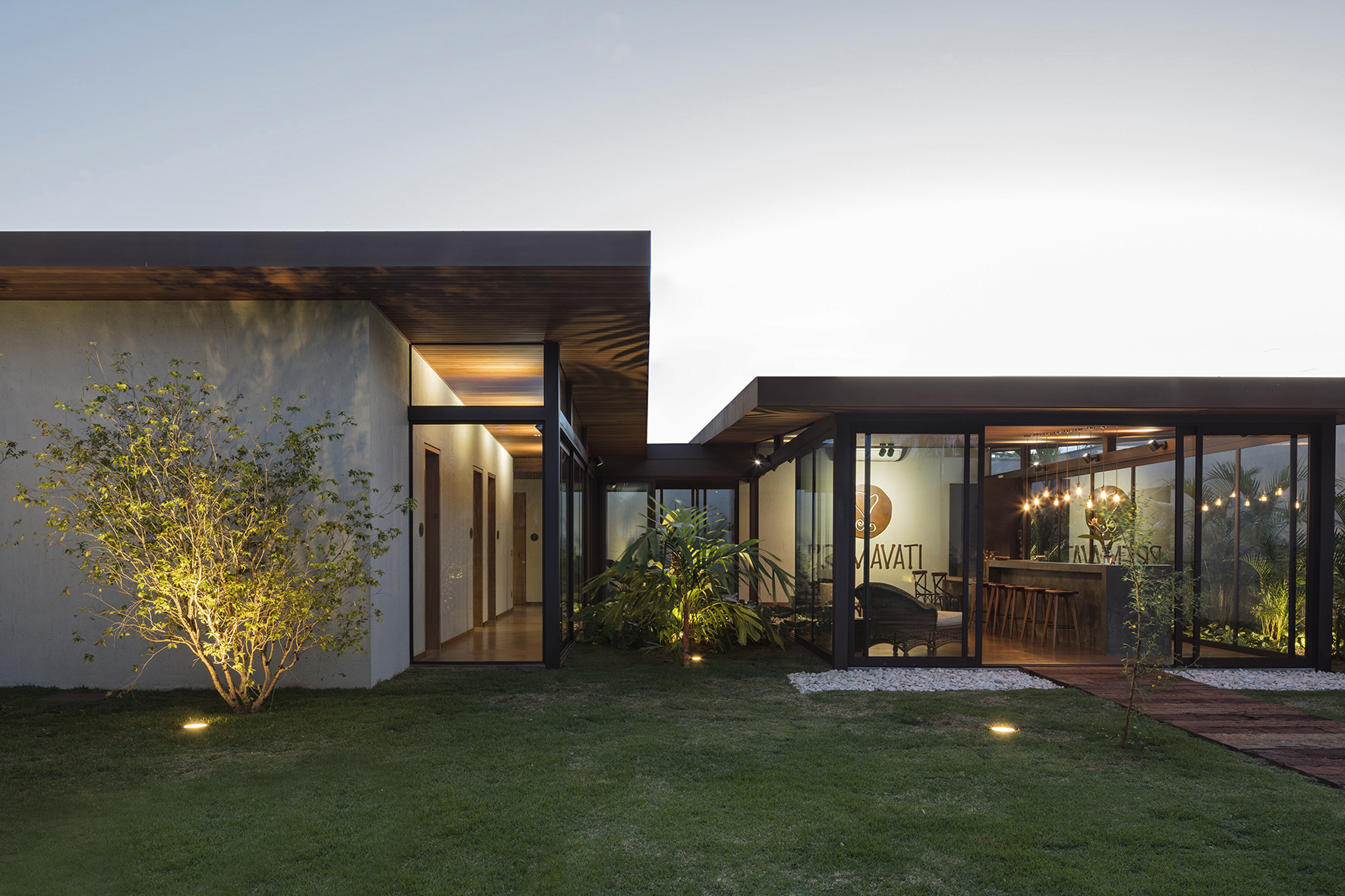 Low pavilion with a deep timber-lined roof overhang and glazed lounge, with a lit shrub on the lawn at twilight