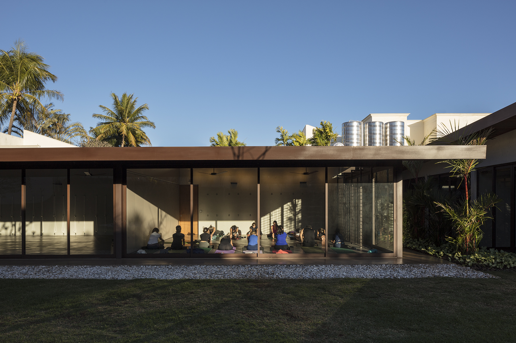 Glazed studio facade with a group seated inside for class, palm trees and water tanks behind in late sun