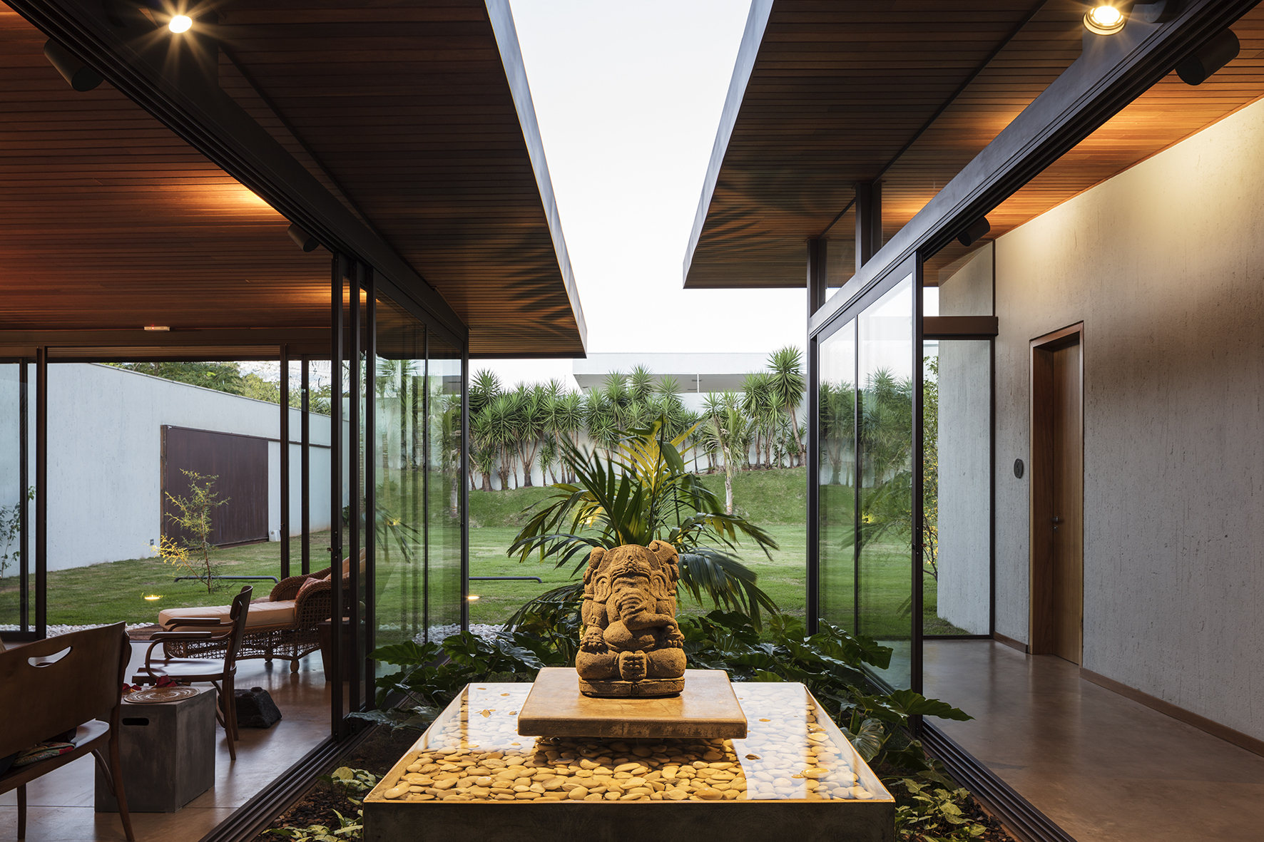 Courtyard water basin with a carved stone Ganesha figure and pebbles, framed by timber soffits and glass walls at dusk