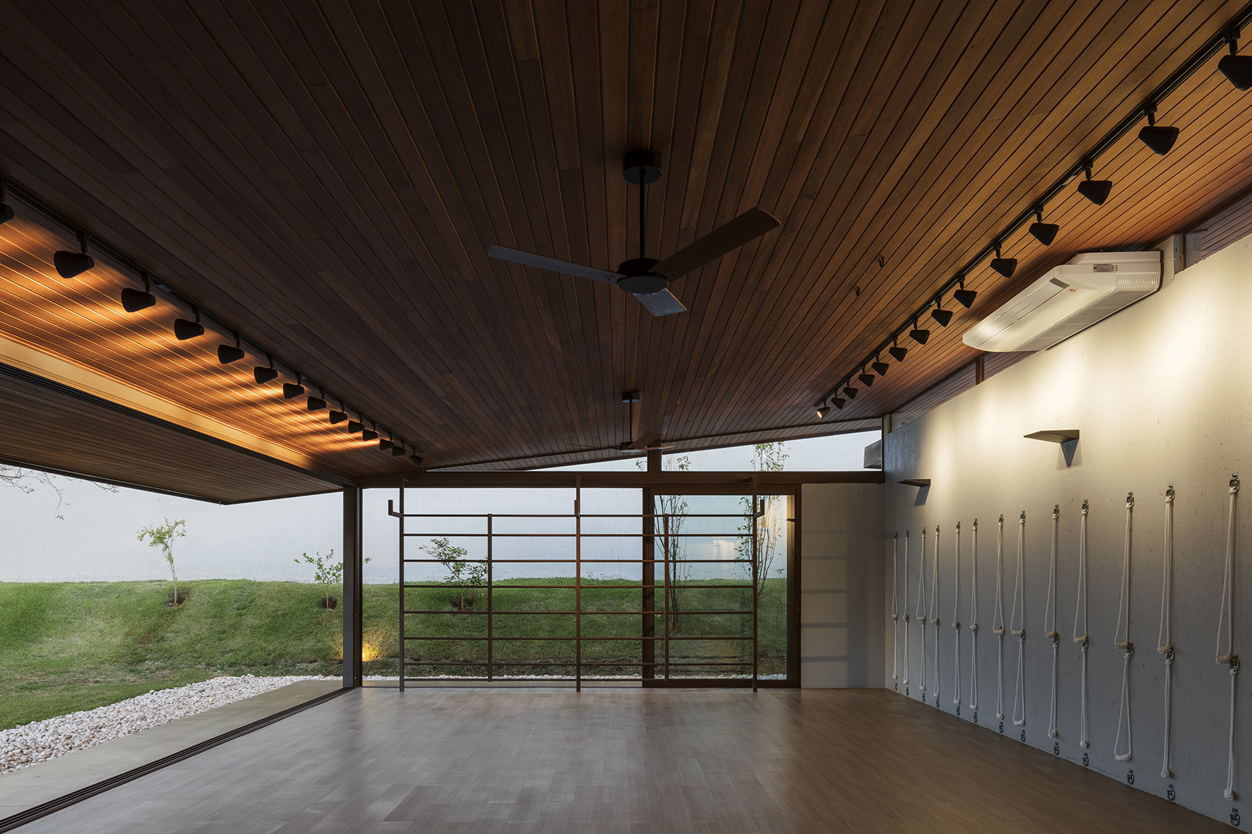 Open studio interior with a slatted timber ceiling, track lights, ceiling fan and rope wall fixtures facing a grassy slope