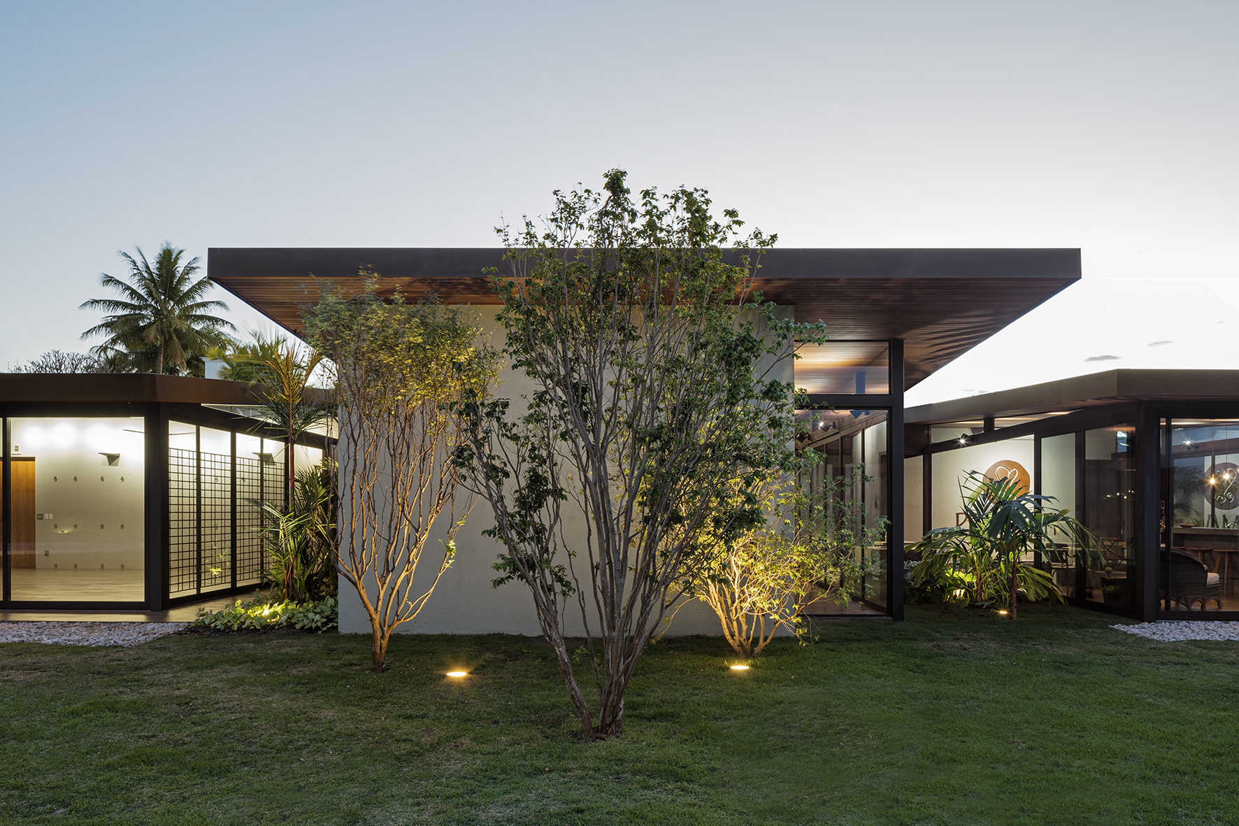Low flat-roofed pavilion with glazed walls behind multi-stemmed trees, ground lights glowing on the lawn at dusk