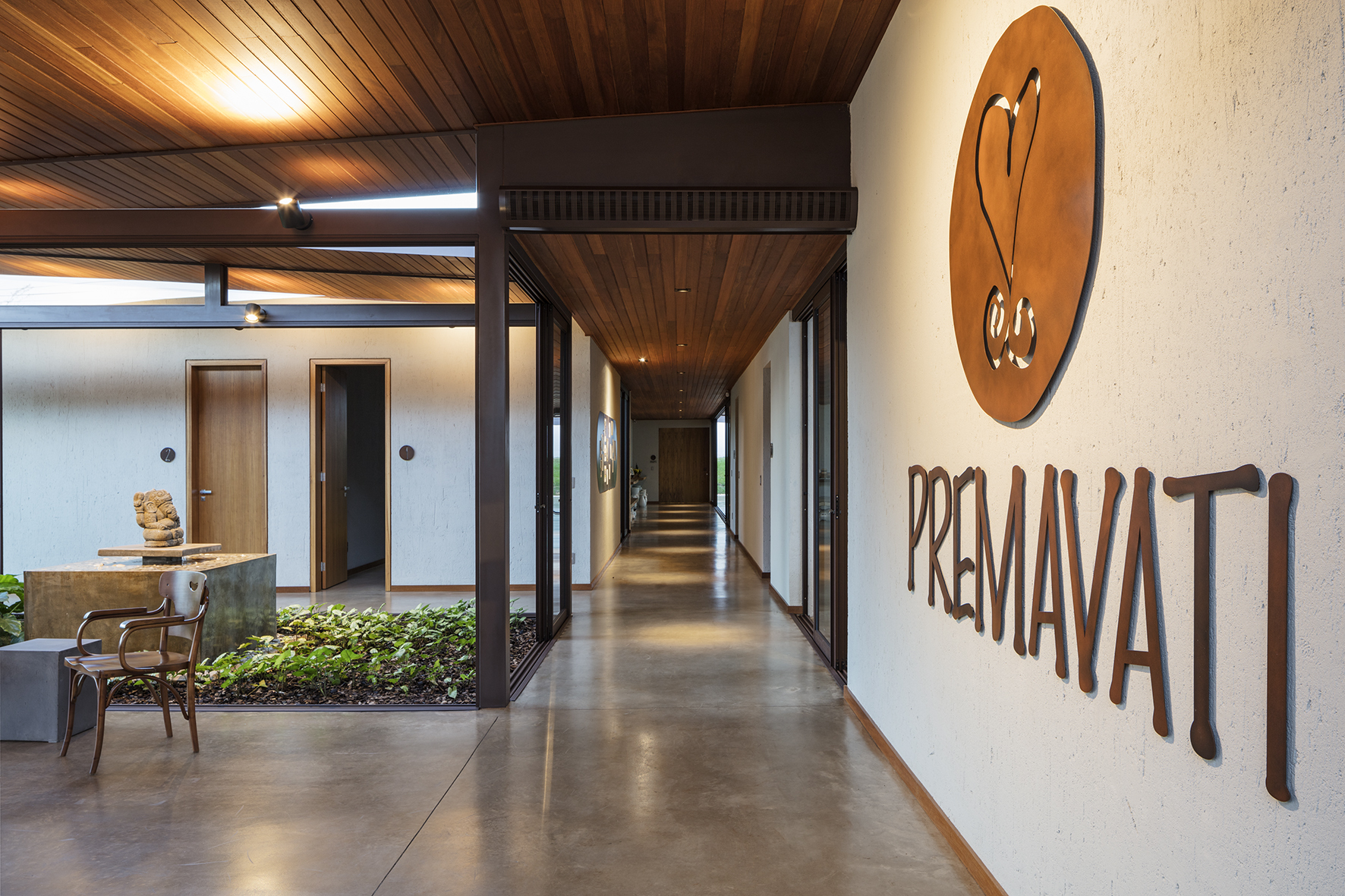 Corridor with timber ceiling and steel columns running past a planted courtyard and a stone sculpture, rust-colored lettering on the wall